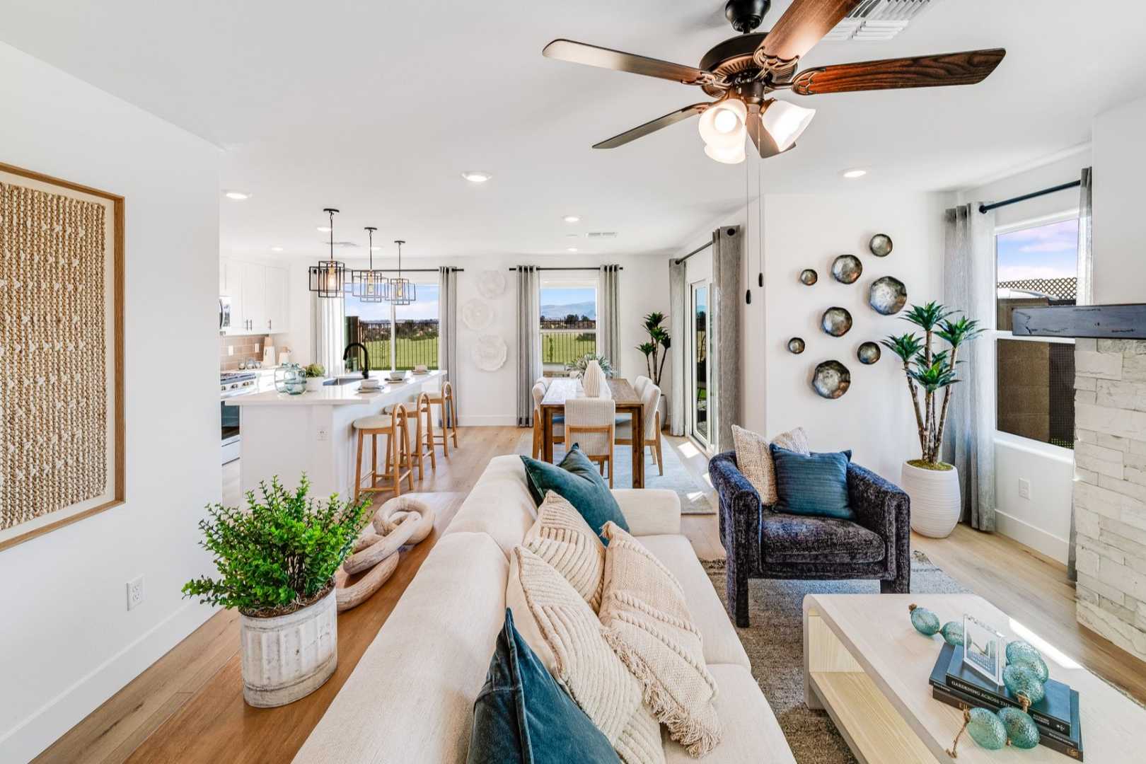 Open-concept living room in The Wilmington: white sofa with teal pillows, stone fireplace, potted plants, kitchen island, large windows