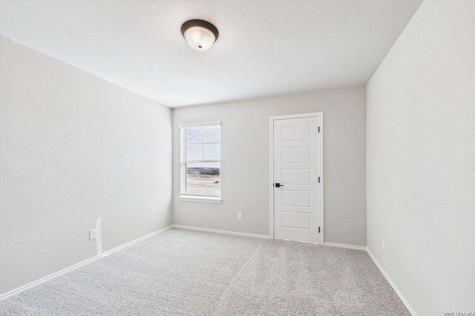 Bright secondary bedroom with light gray walls, large window, white door, and carpet floor in Davidson Homes The Charlotte A, San Antonio