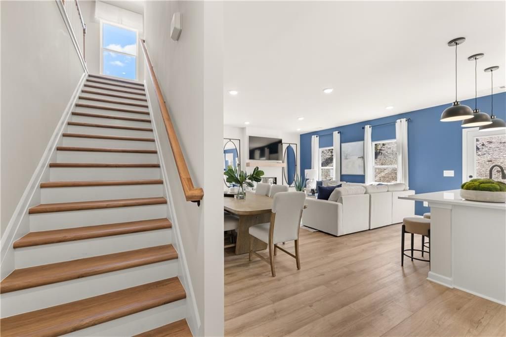 Elegant wooden staircase to sunlit window beside open living-dining area, blue walls, modern sofa, kitchen island in The Burton C by Davidson Homes, Canton, GA
