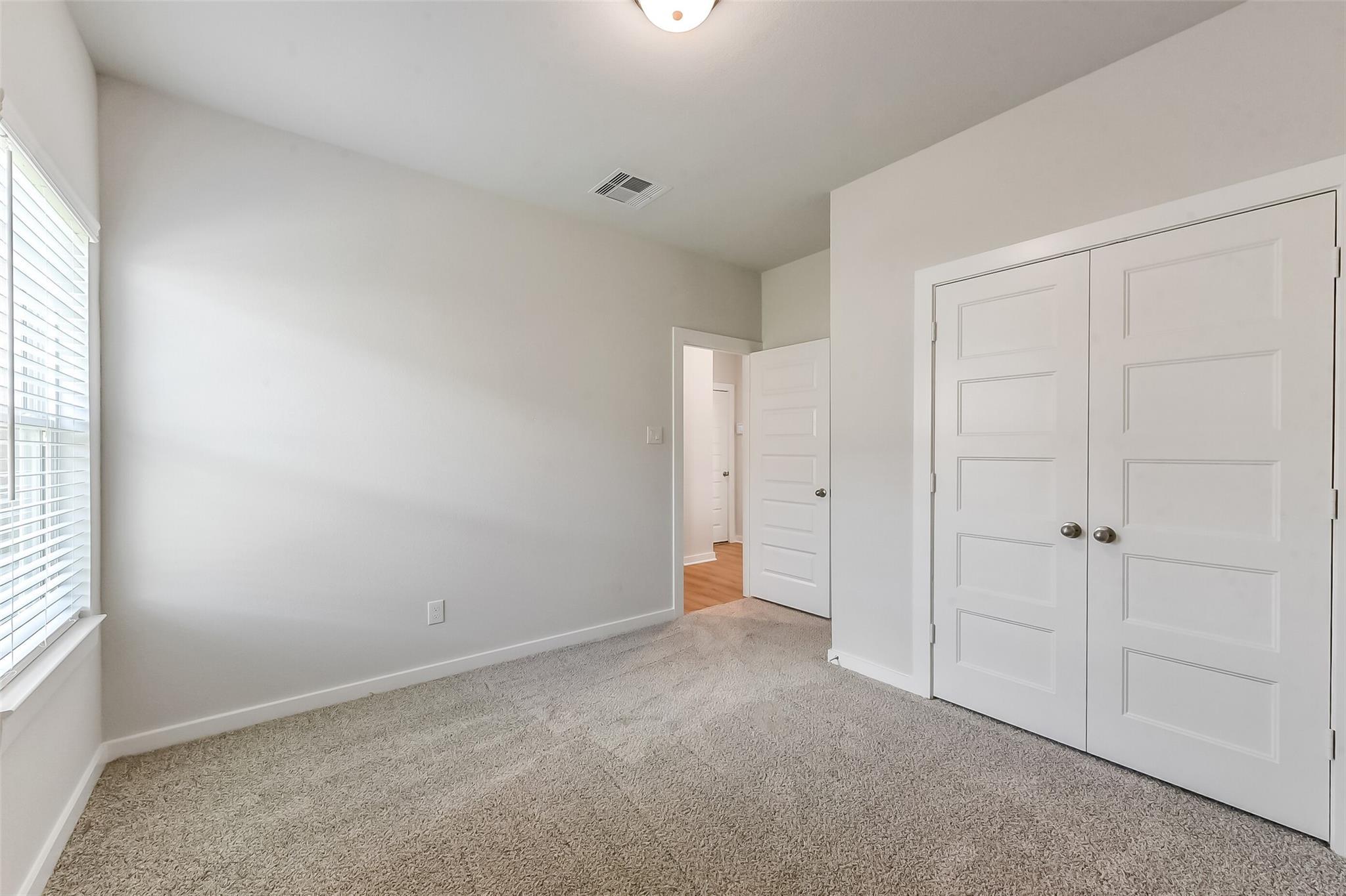 Empty bedroom with gray carpet, white walls, window blinds, double closet doors in Davidson Homes The Costa C, Dayton, Texas