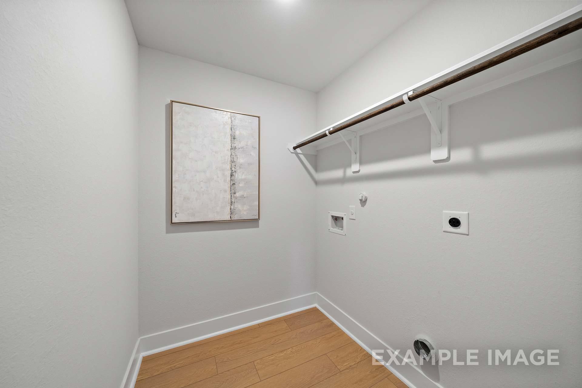 Spacious laundry room in The Rose B home by Davidson Homes with white shelving, hanging rod, washer-dryer hookups, abstract wall art, and hardwood floors