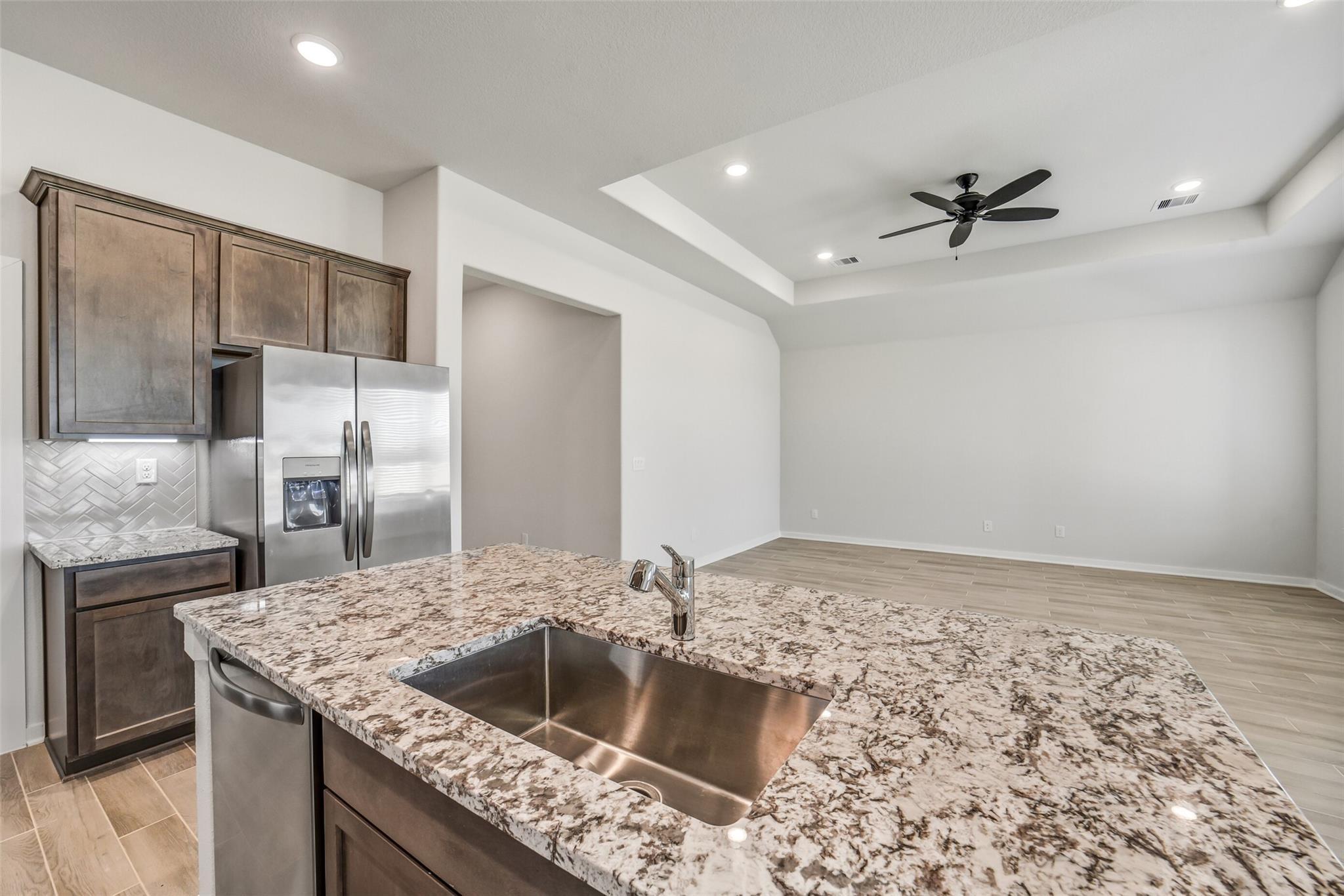 Modern kitchen with granite island, stainless fridge, brown cabinets, herringbone backsplash in Davidson Homes Acadia C, Magnolia TX