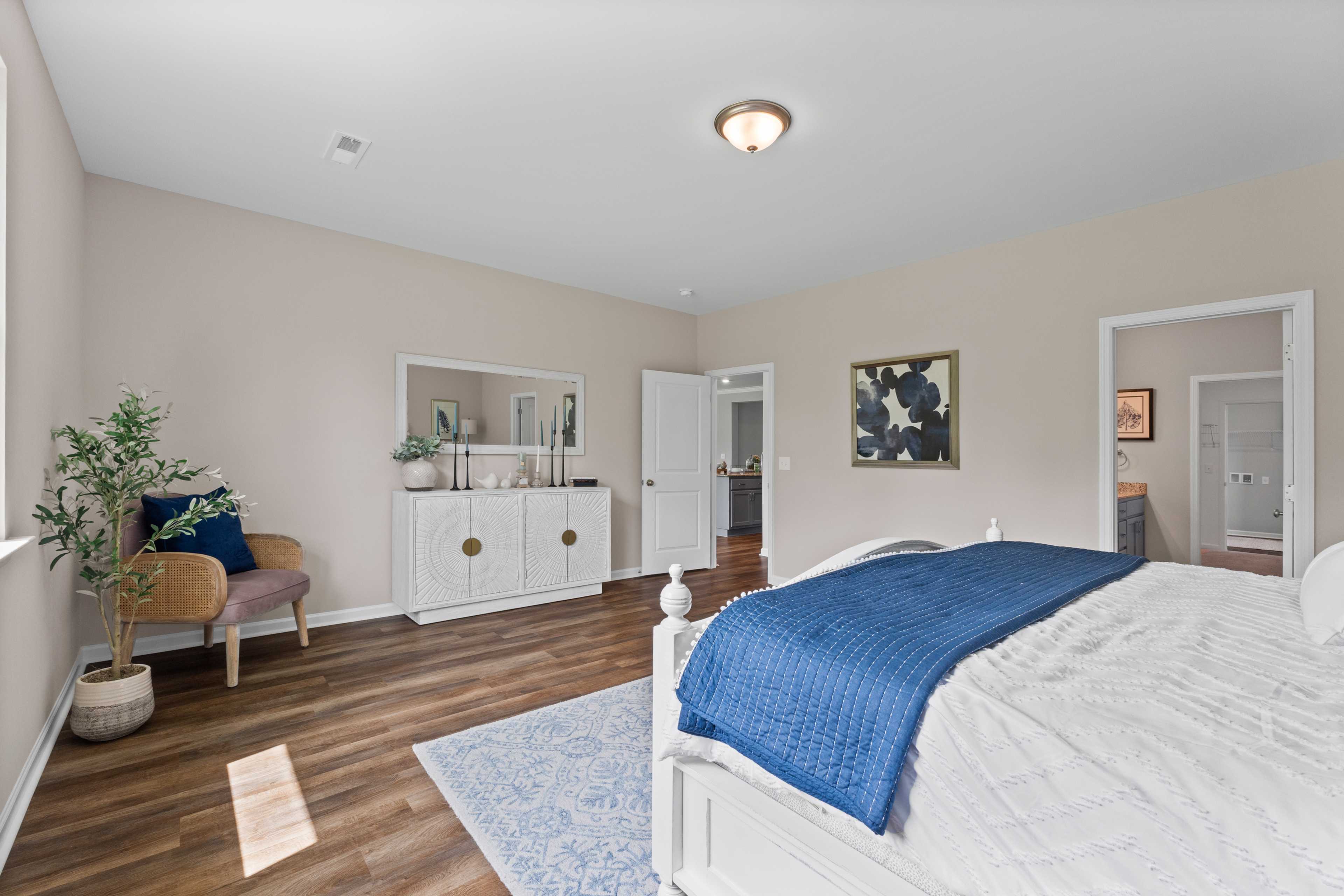 Spacious primary bedroom at Riverton Preserve in Huntsville Alabama with hardwood floors, white bed, blue quilt, accent chair, and potted plant