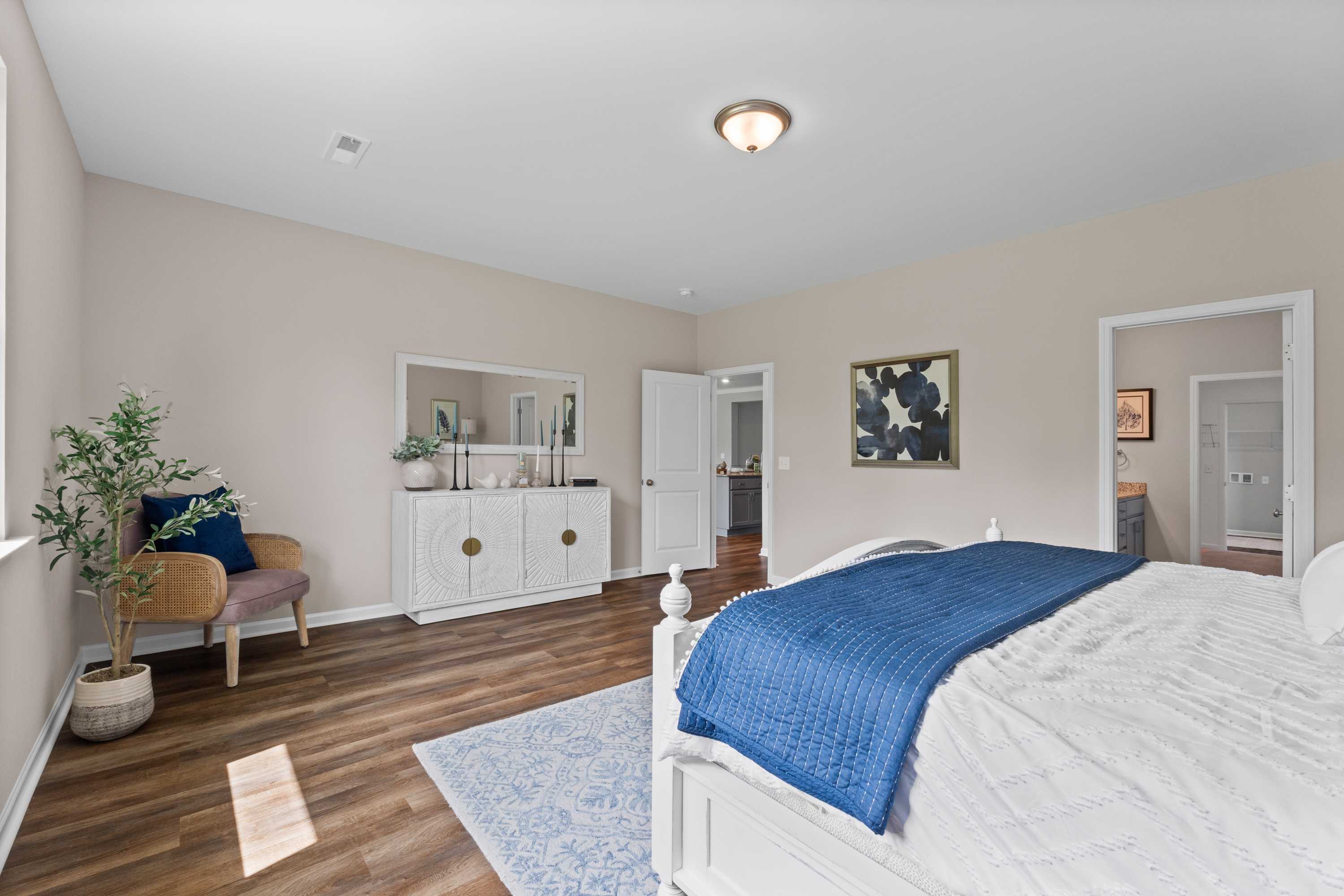 Spacious primary bedroom at Riverton Preserve in Huntsville Alabama with hardwood floors, white bed, blue quilt, accent chair, and potted plant