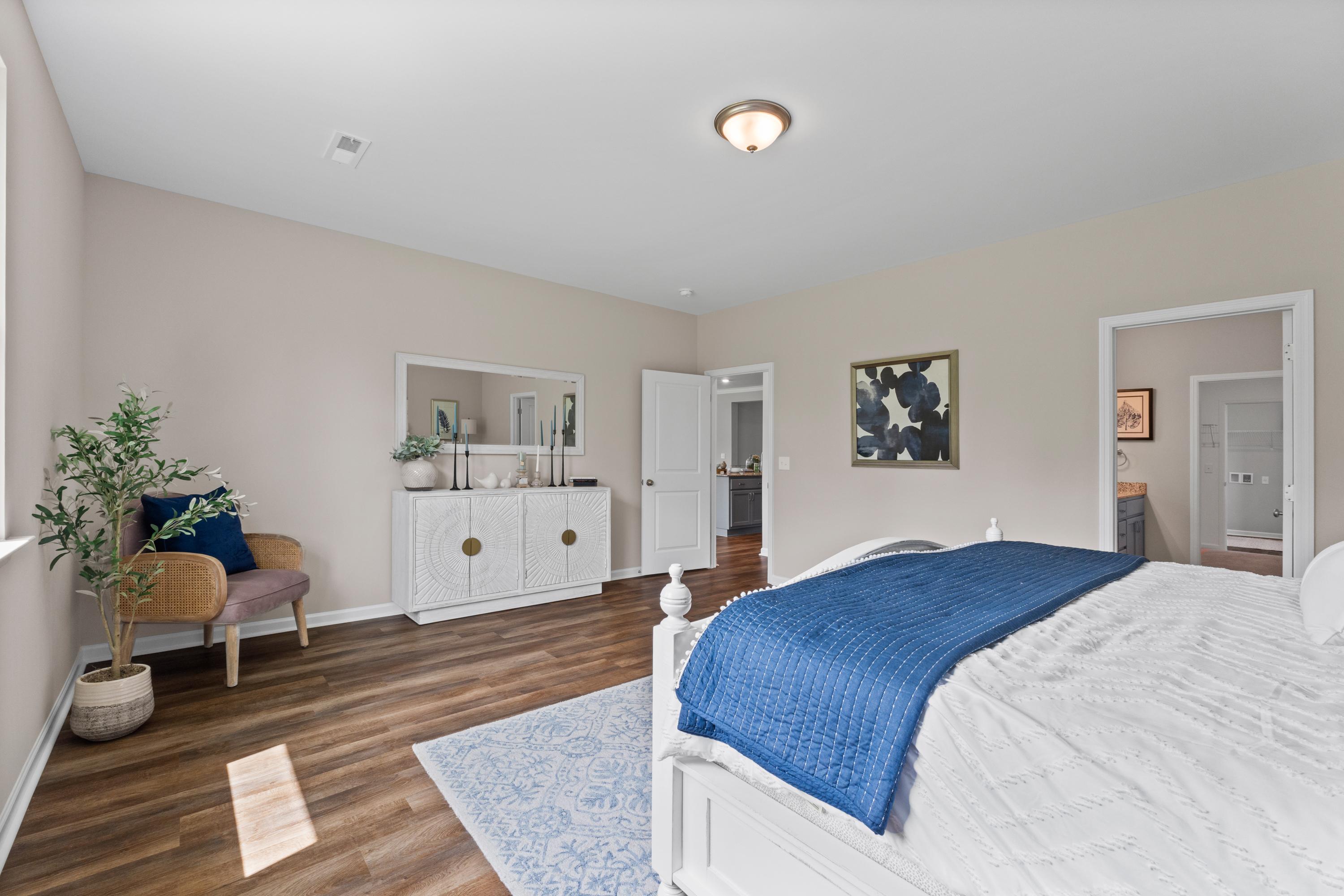 Spacious primary bedroom at Riverton Preserve in Huntsville Alabama with hardwood floors, white bed, blue quilt, accent chair, and potted plant