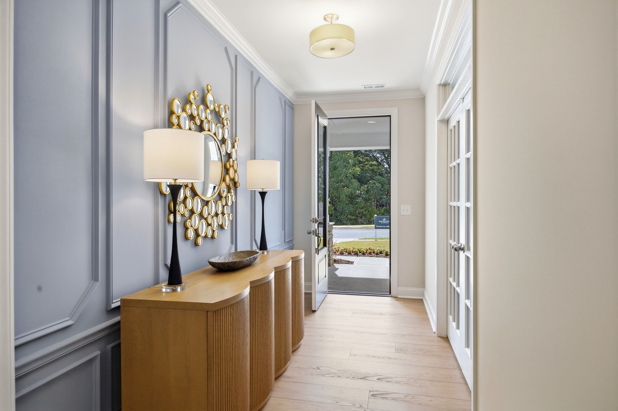 Elegant foyer hallway at Enclave at Belmont in Belmont NC with blue paneled walls, gold mirror, wooden consoles, hardwood floors, and open door to lush landscaping
