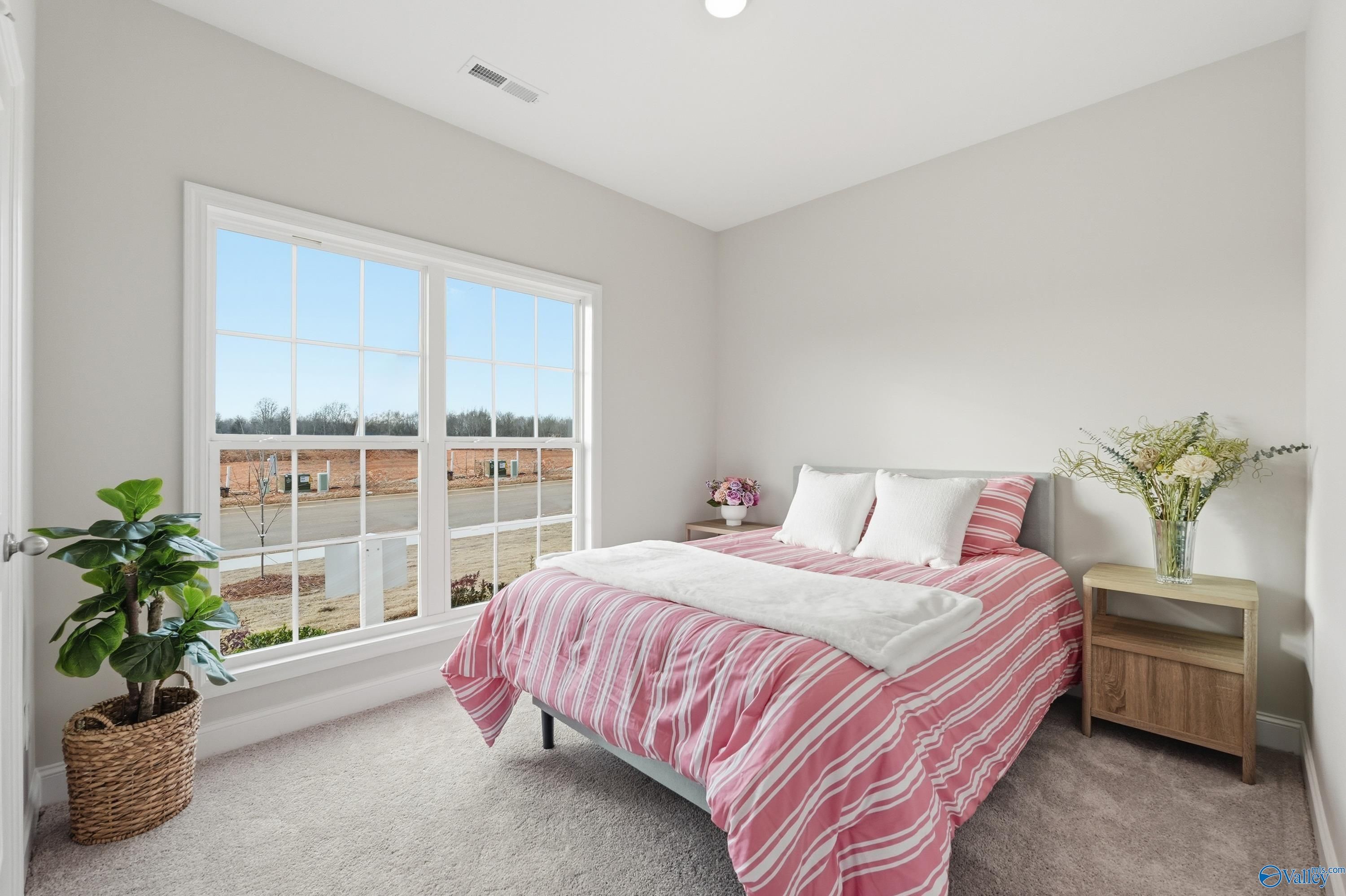 Cozy bedroom with pink striped bedding, white pillows, floral vase on nightstand, potted plant, and large window in Davidson Homes The Asheville, Toney, Alabama