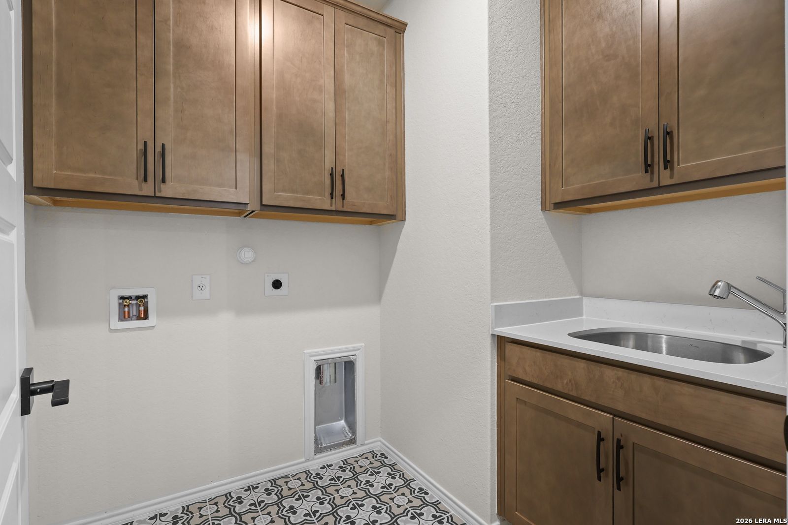 Laundry room with wood cabinets, stainless sink, quartz countertop, and patterned tile floor in Davidson Homes Glenwood E, San Antonio