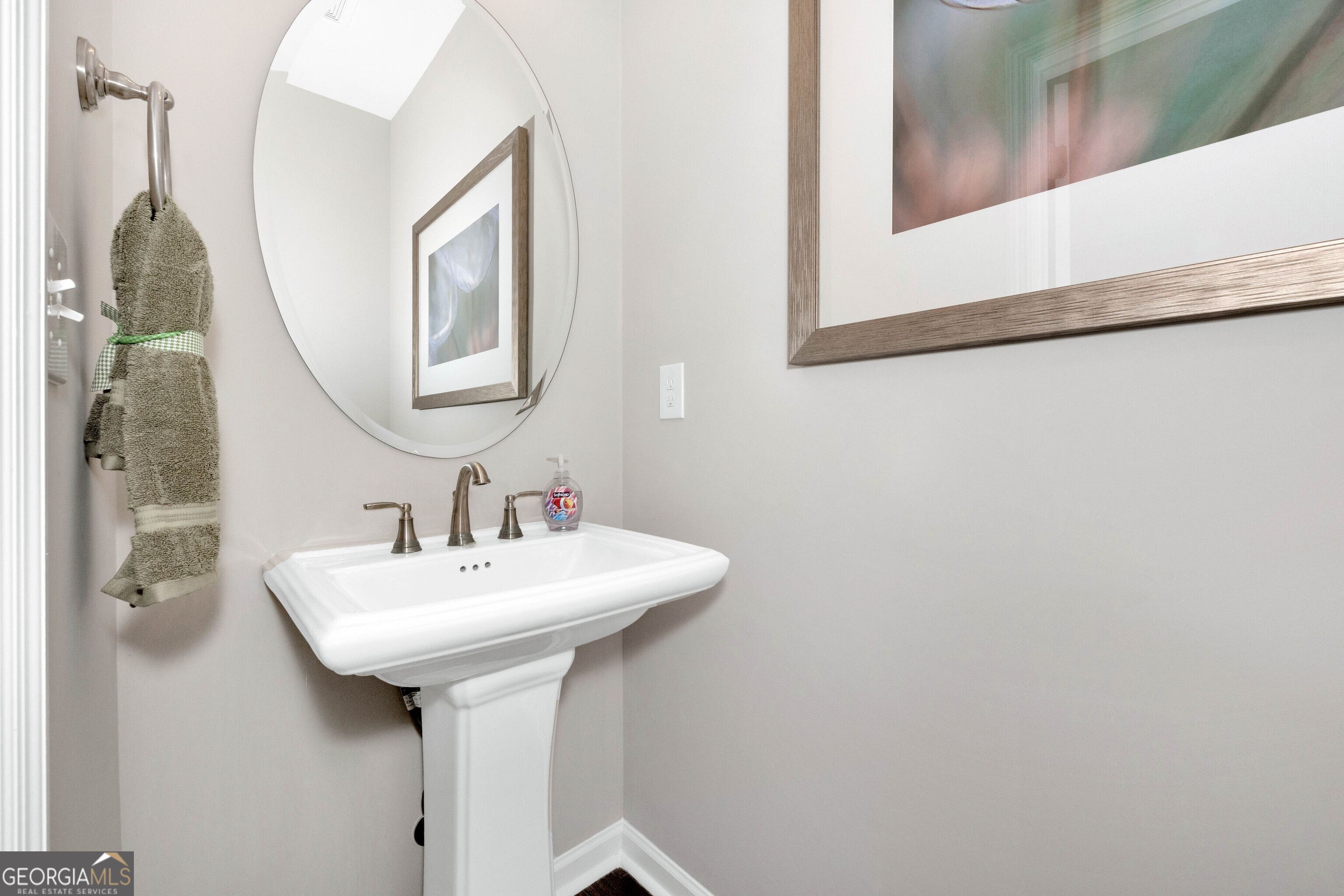 Elegant powder room with white pedestal sink, round mirror, green towel, and abstract art in Evermore Homes The Stella, Ivy Glen, Perry, Georgia