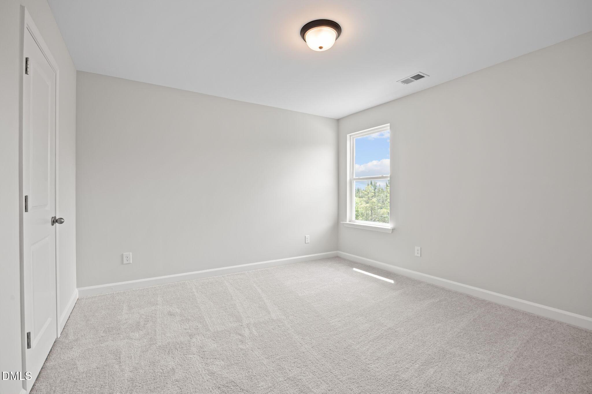 Bright empty bedroom with neutral gray walls, plush carpet, ceiling light, and window view of trees in Davidson Homes The Willow D, Wendell, NC