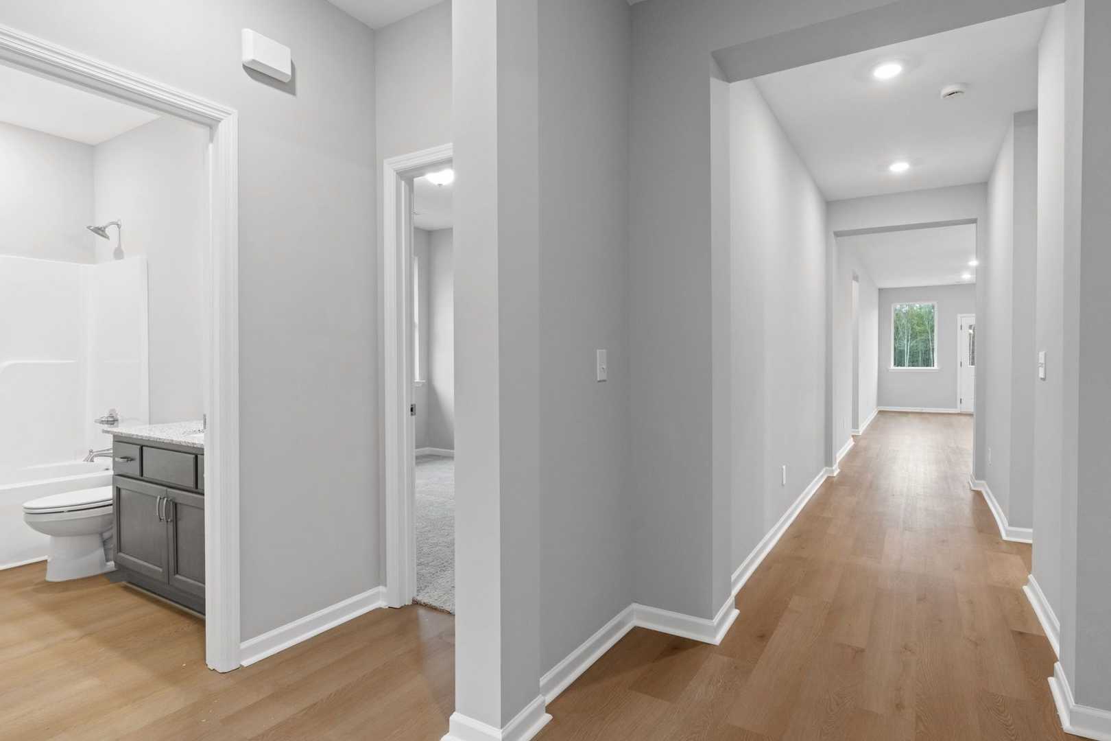 The Luna master bathroom featuring white tub, walk-in shower, gray vanity, and adjacent hallway with hardwood floors
