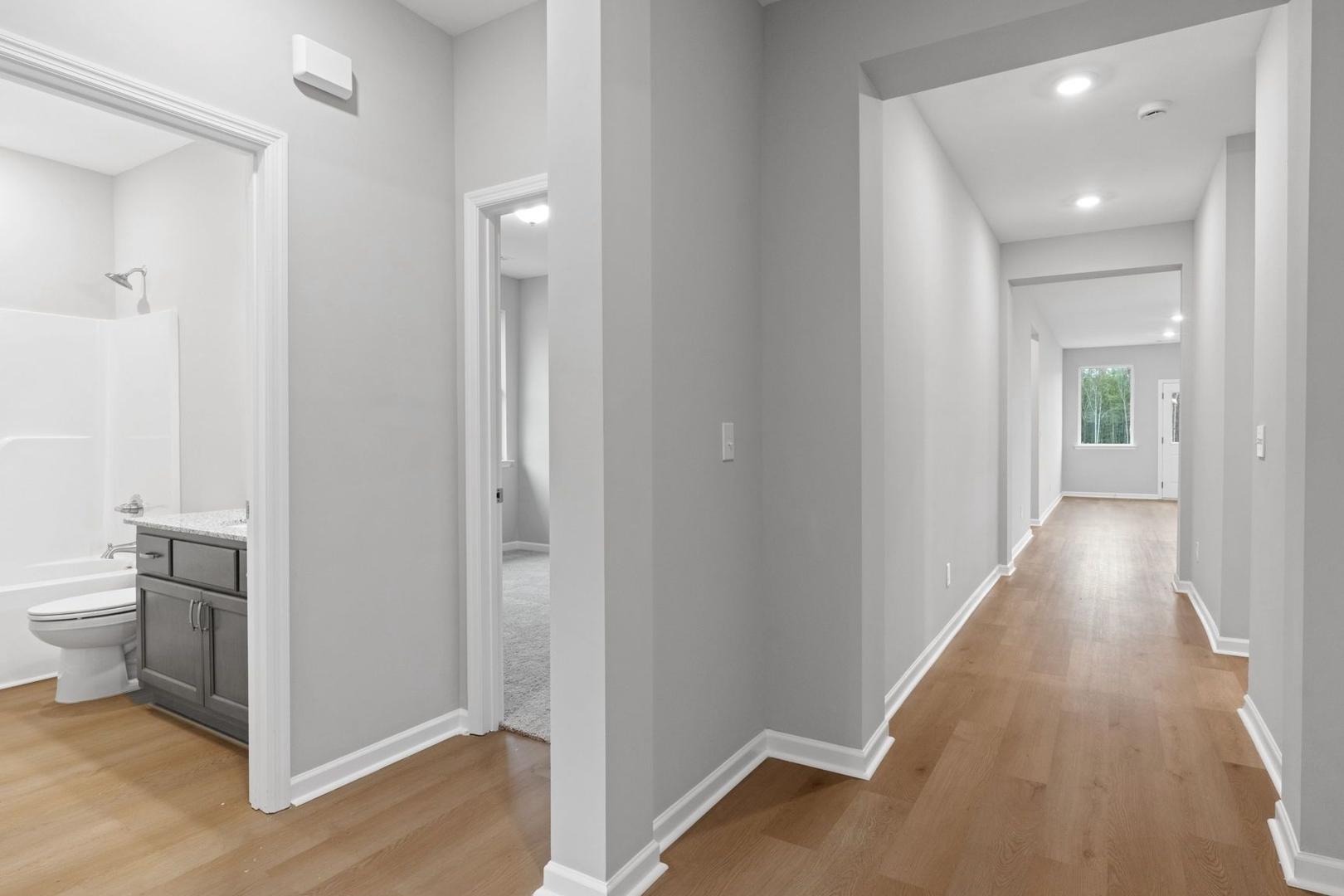 The Luna master bathroom featuring white tub, walk-in shower, gray vanity, and adjacent hallway with hardwood floors