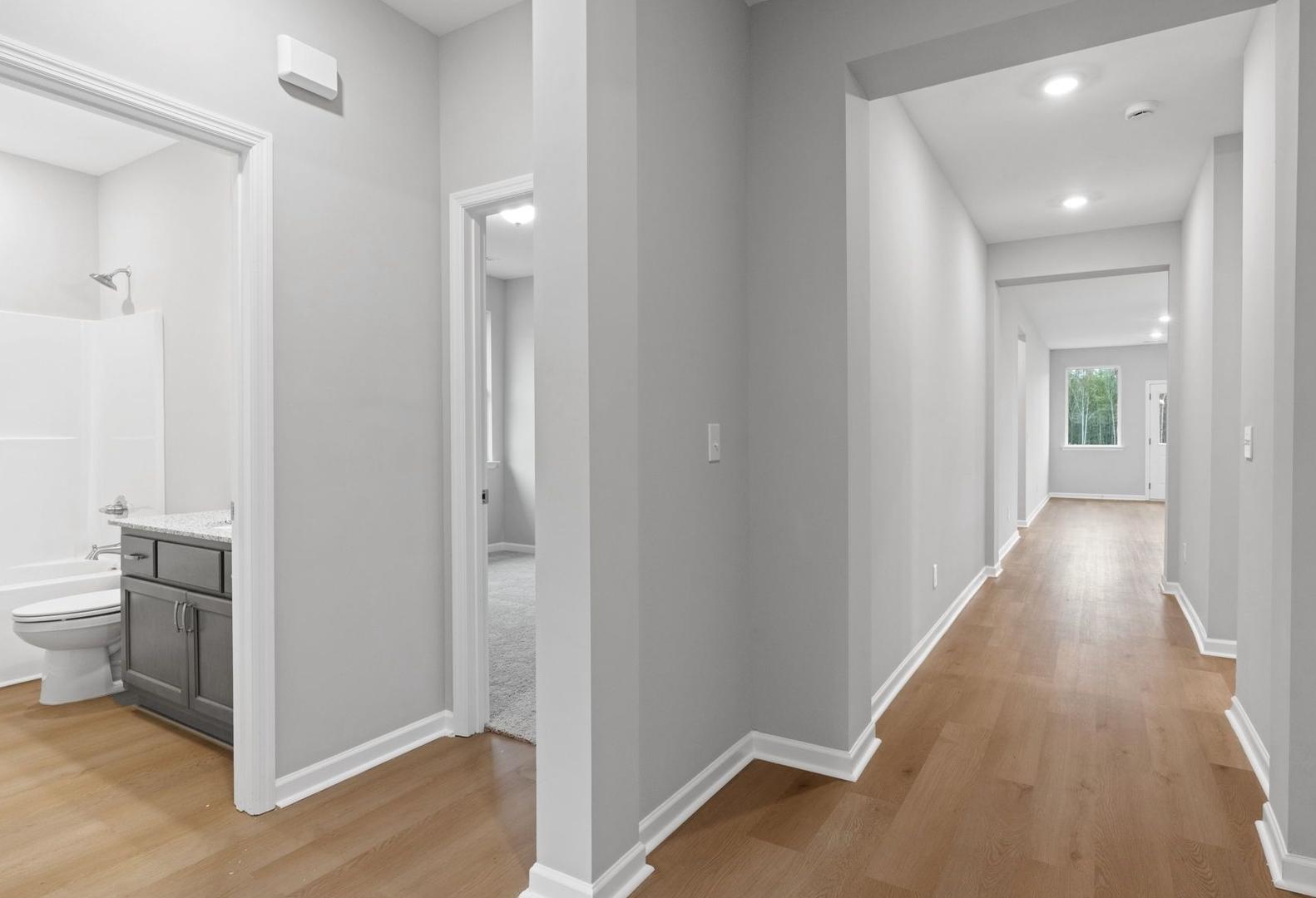 The Luna master bathroom featuring white tub, walk-in shower, gray vanity, and adjacent hallway with hardwood floors