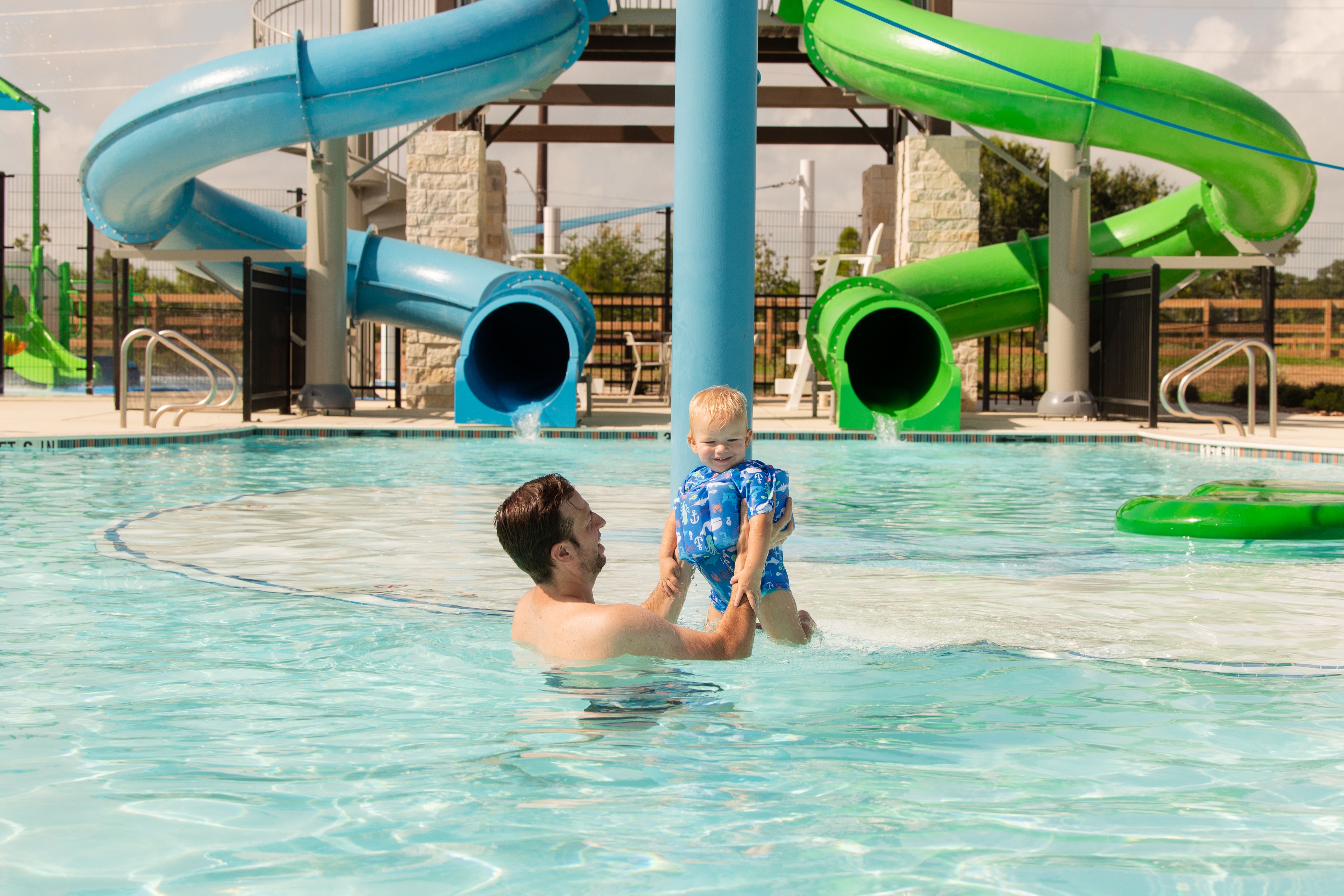 Father holding toddler in resort-style pool with blue and green twisting water slides at Emberly in Beasley, Texas