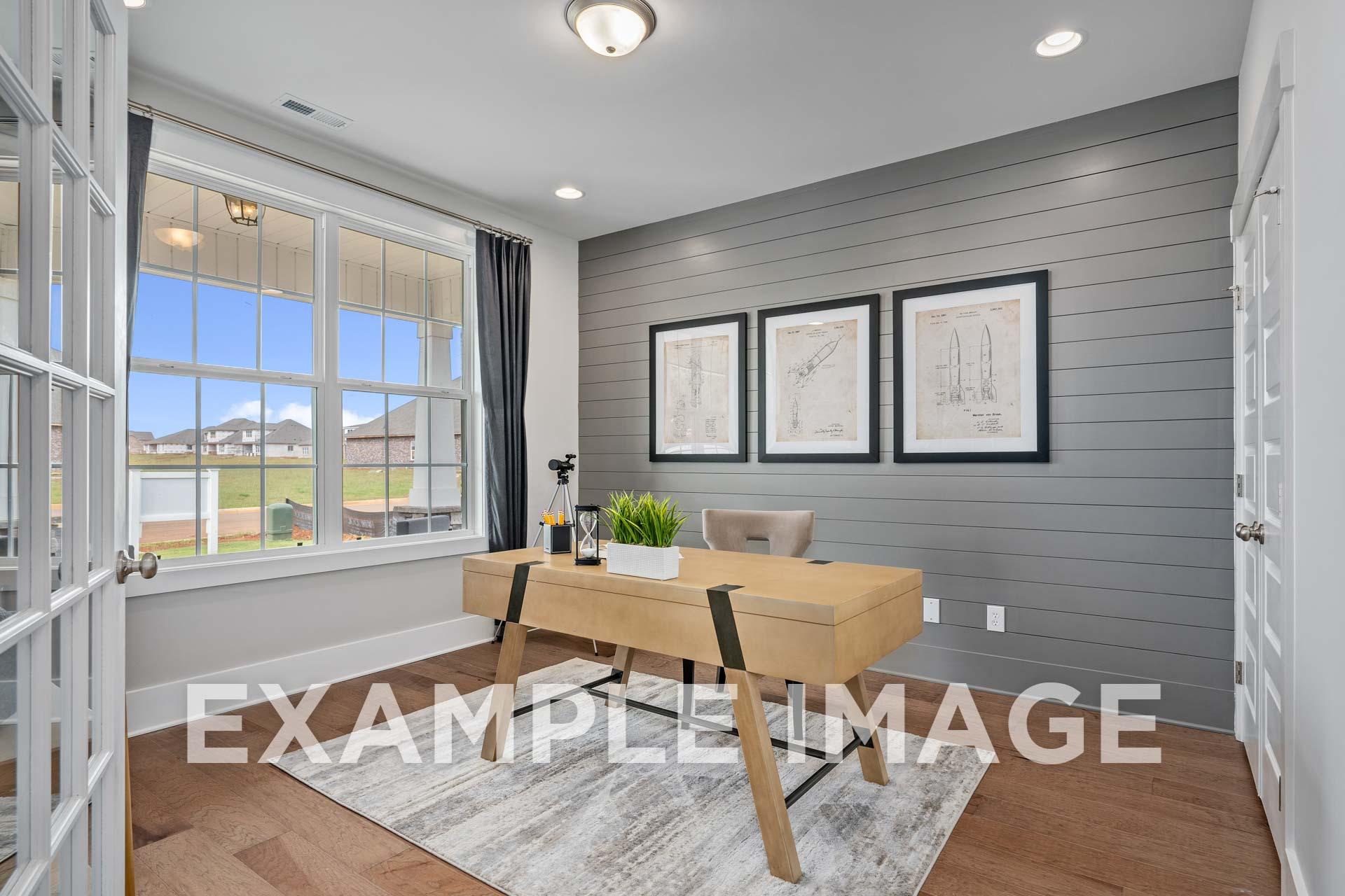Spacious home office in The Harrison with wooden desk, blueprint art on gray shiplap walls, and large windows to green view