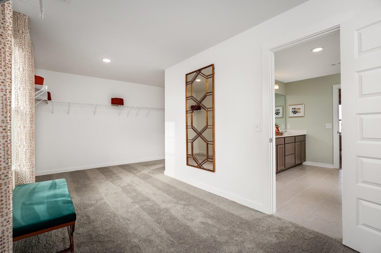 Elegant walk-in closet with gold geometric mirror, built-in shelving, and green bench opening to sage green master bathroom in Davidson Homes The Gordon C, White House TN