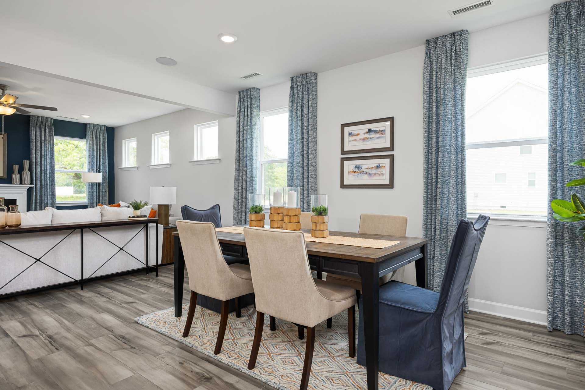 Open-concept dining room at Beverly Place in Four Oaks NC with hardwood floors, beige upholstered chairs, wooden table, and blue curtains