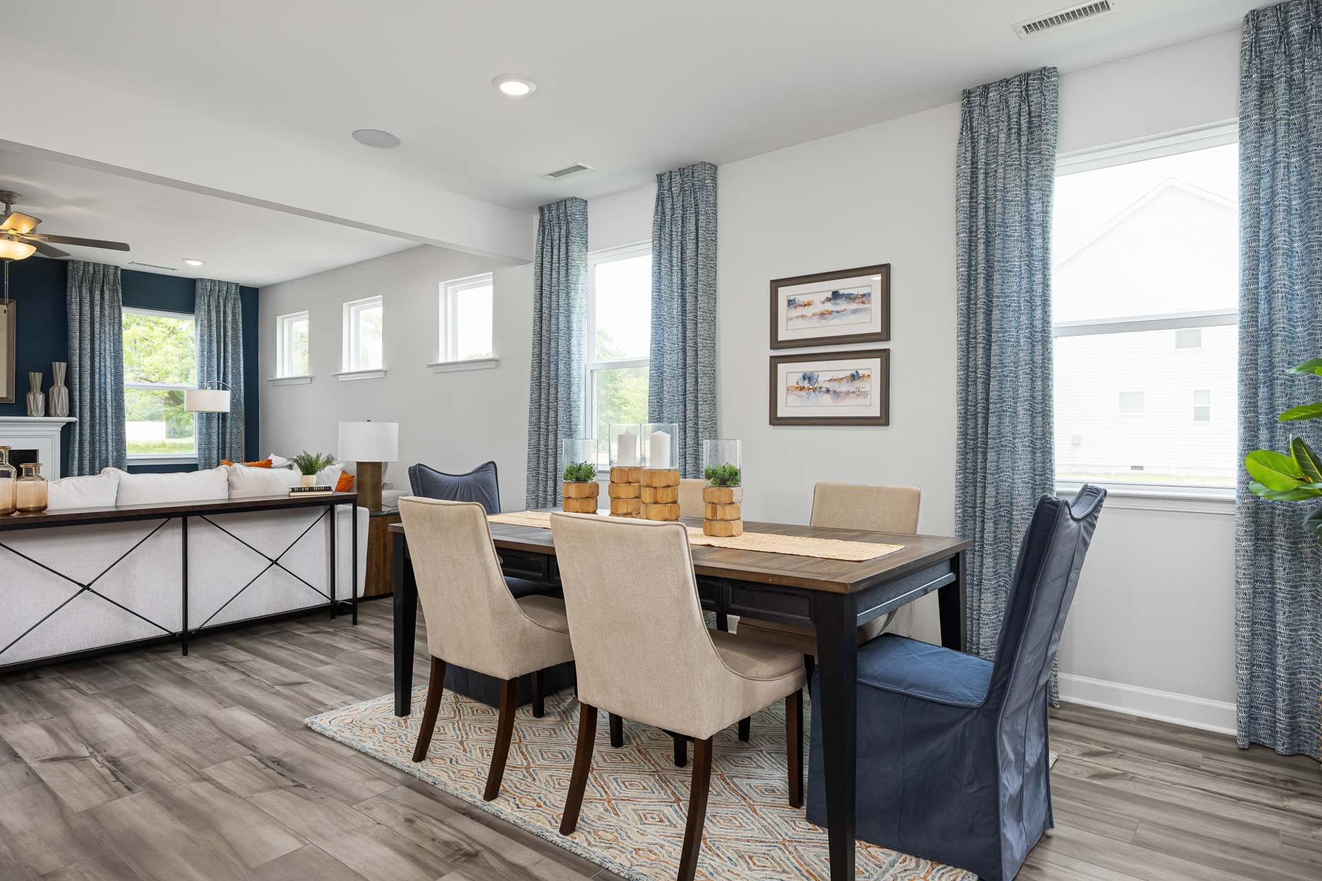 Open-concept dining room at Beverly Place in Four Oaks NC with hardwood floors, beige upholstered chairs, wooden table, and blue curtains
