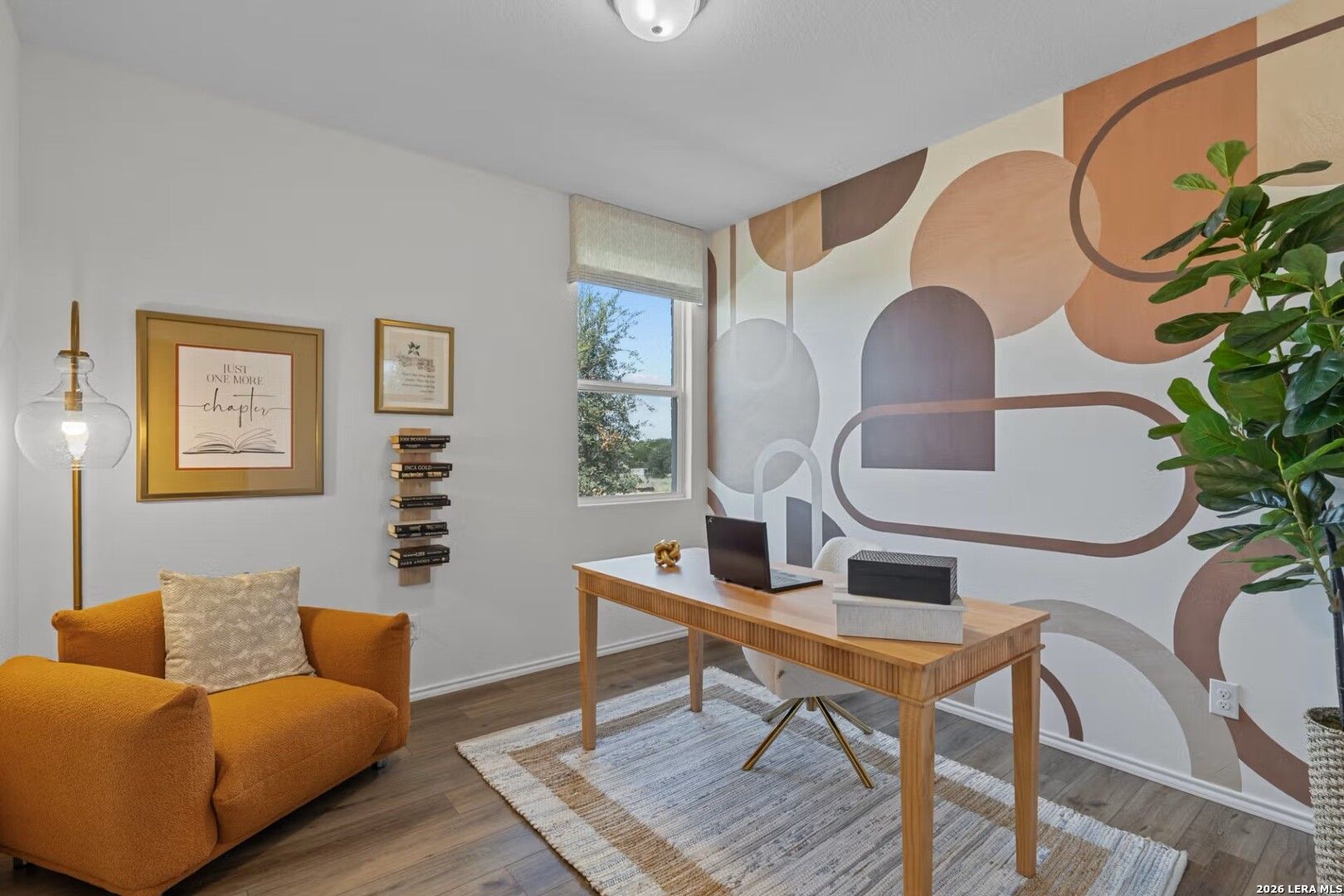 Modern home office with wooden desk, laptop, orange armchair, abstract beige wall art, and potted plant in The Daphne K, San Antonio