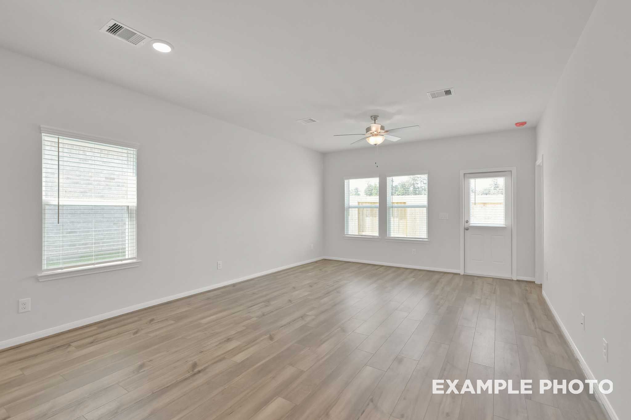 Spacious empty living room in The Comal floor plan with hardwood floors, gray walls, large windows, blinds, and ceiling fan by Davidson Homes