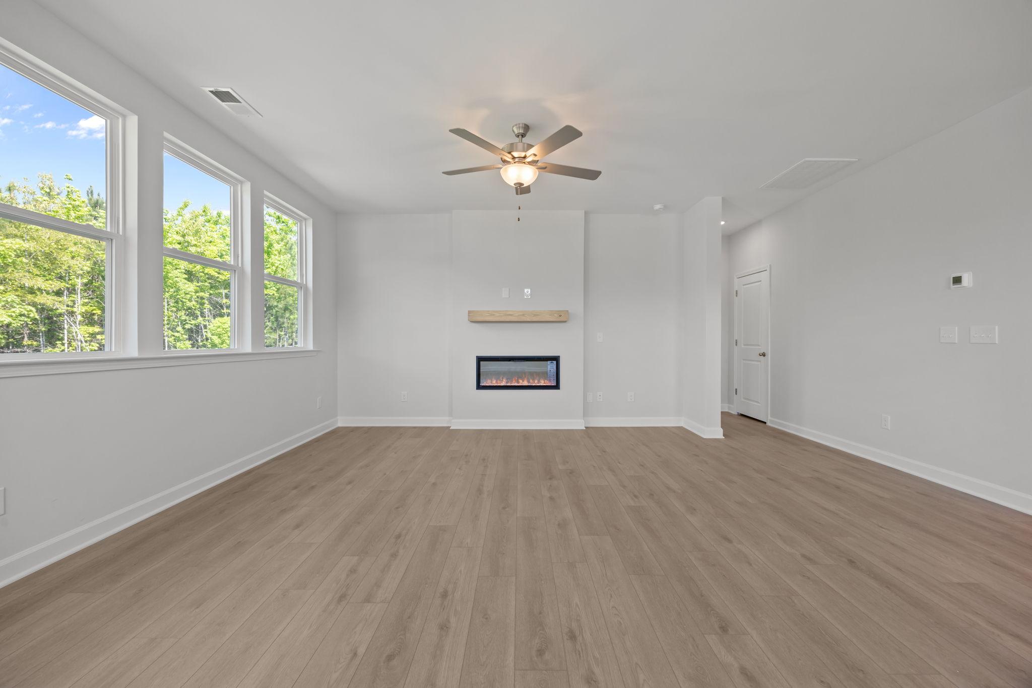 Bright, open-concept living area in The Ashport B featuring a modern electric fireplace, wood-look floors, and large windows.