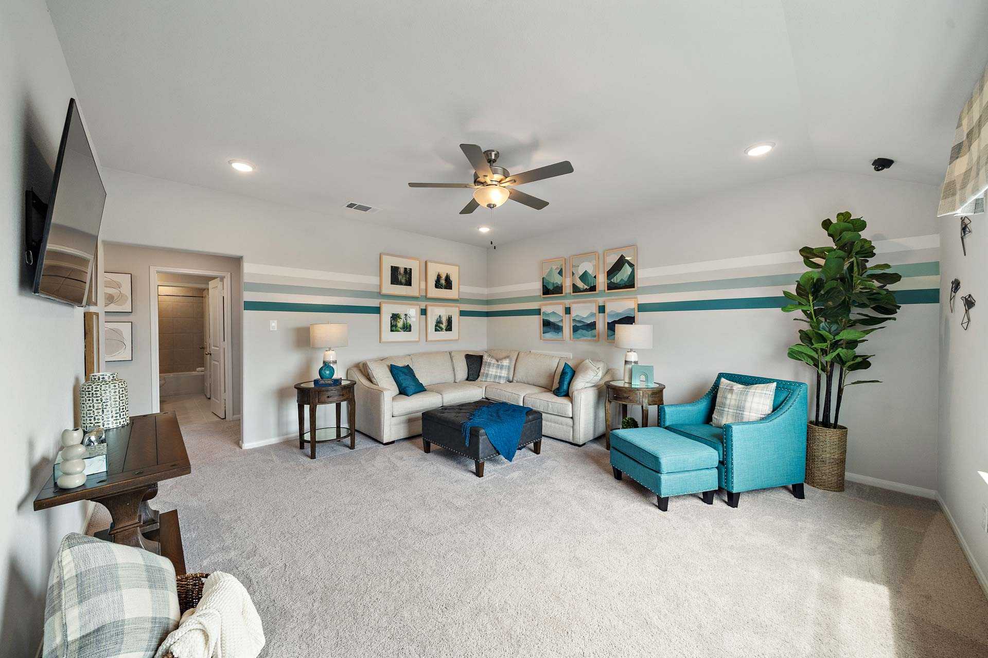Spacious living room in Sunterra Katy Texas home by Davidson Homes featuring L-shaped beige sofa, turquoise accent chair, mounted TV, ceiling fan, and fiddle leaf fig plant
