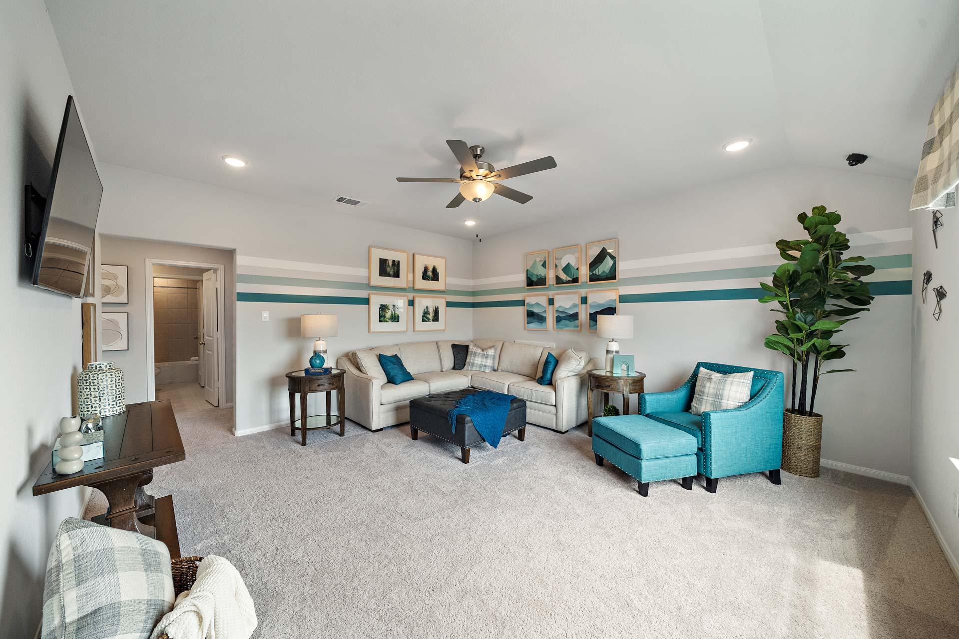 Spacious living room in Sunterra Katy Texas home by Davidson Homes featuring L-shaped beige sofa, turquoise accent chair, mounted TV, ceiling fan, and fiddle leaf fig plant