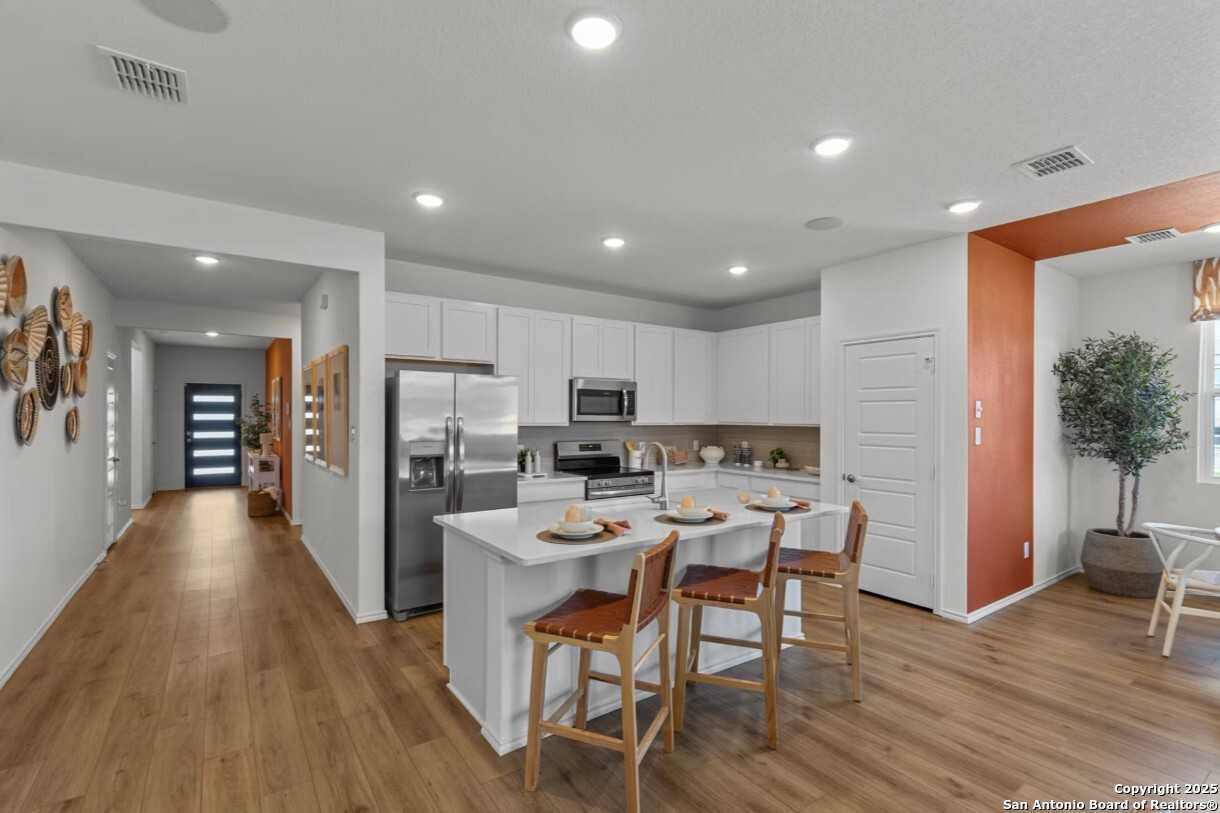 Modern white kitchen with stainless appliances, island bar stools, and open hallway in Davidson Homes The Daphne I, Bricewood, San Antonio