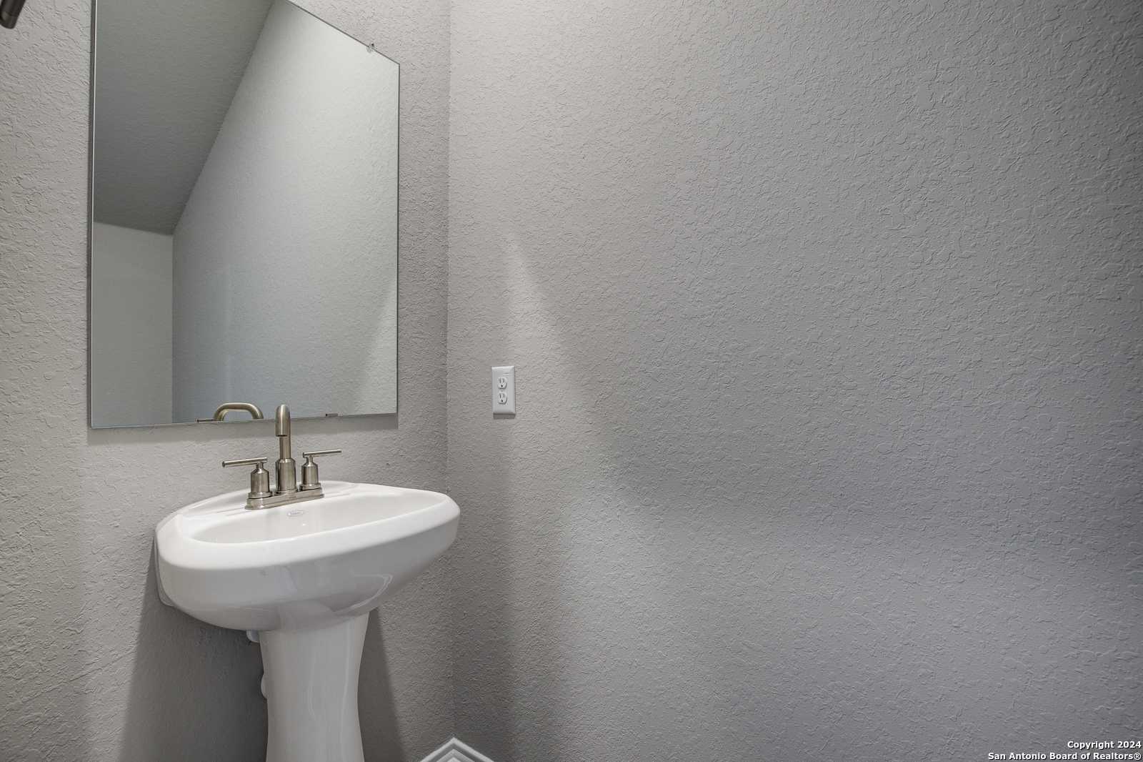 Elegant powder room with white pedestal sink, brushed nickel faucet, and frameless mirror on neutral gray walls in Davidson Homes The Blanco C, San Antonio