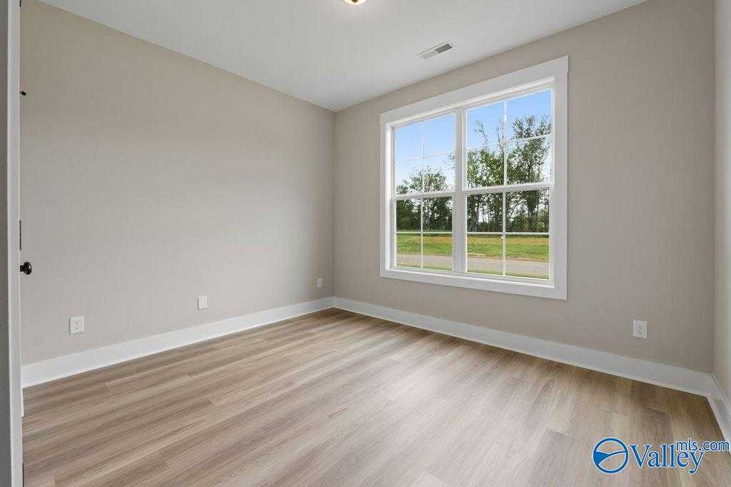 Bright empty bedroom with beige walls, large window overlooking green lawn, and light wood floors in Evermore Homes The Oxford, Owens Cross Roads, Alabama