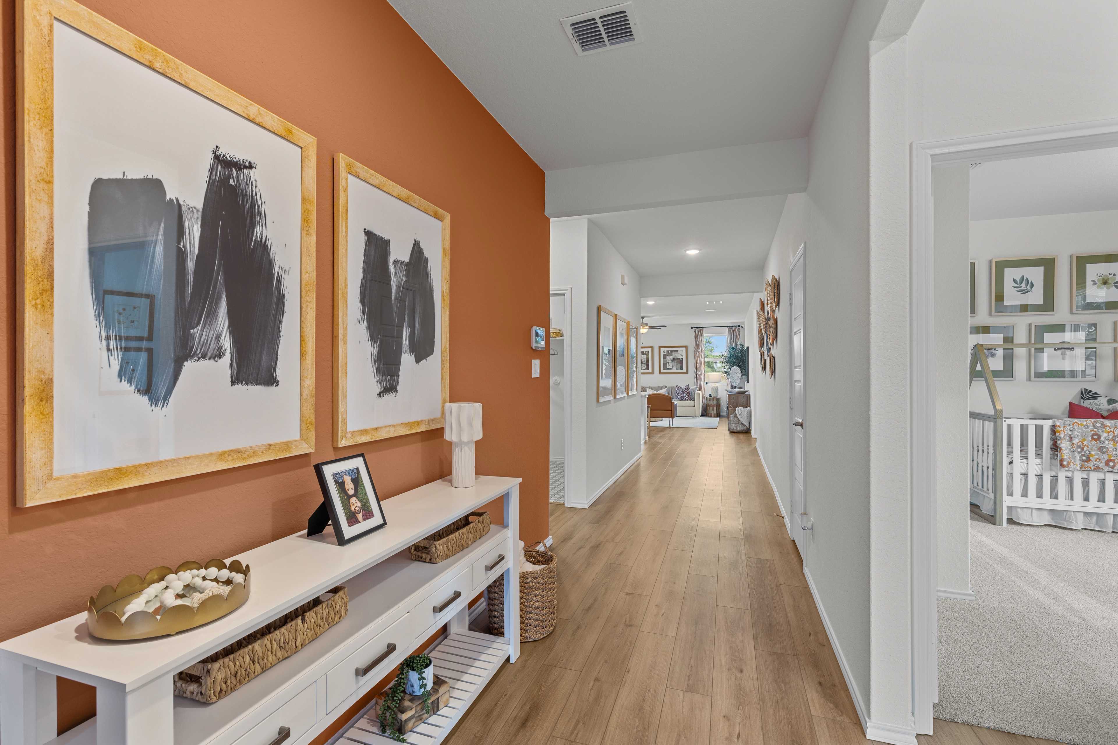 Spacious hallway in The Daphne H with burnt orange accent wall, abstract black art frames, white console table, and wood floors leading to nursery