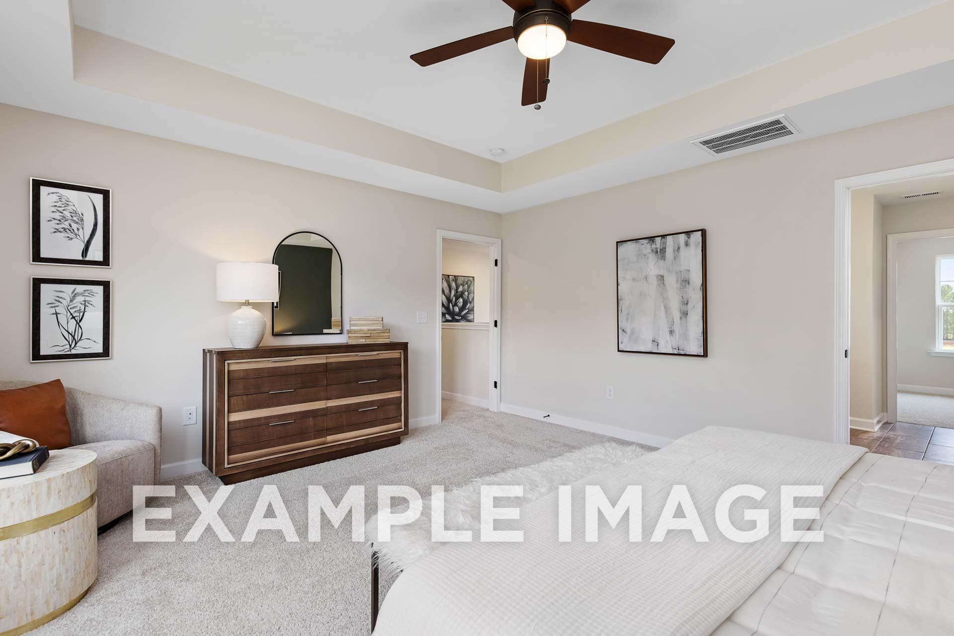 Spacious master bedroom in The Willow E featuring beige walls, ceiling fan, wooden dresser, arched mirror, abstract art, and king bed