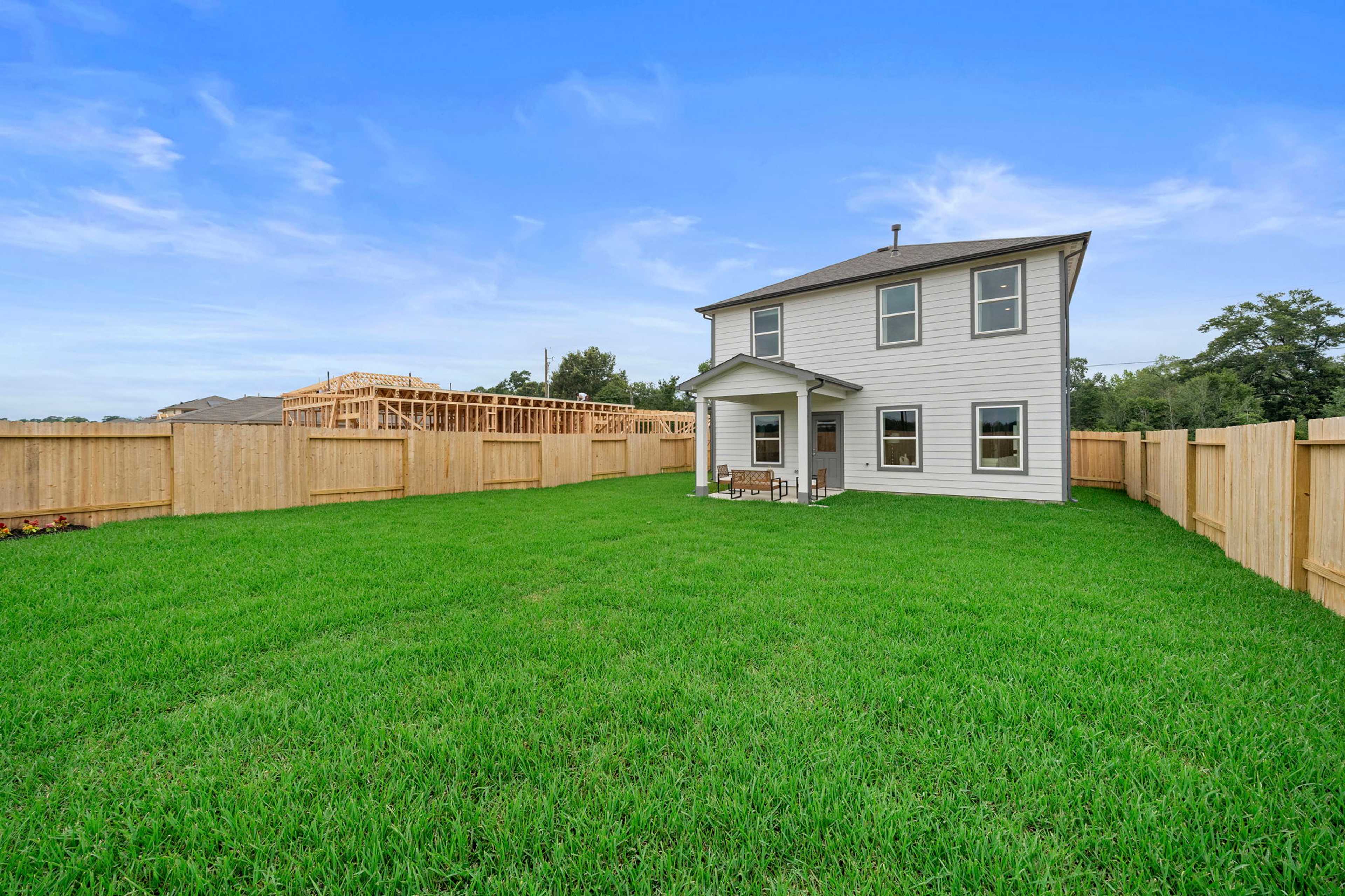 Modern white home with covered porch at Spring Branch Crossing in Conroe TX, lush green yard, wooden fence, and construction backdrop