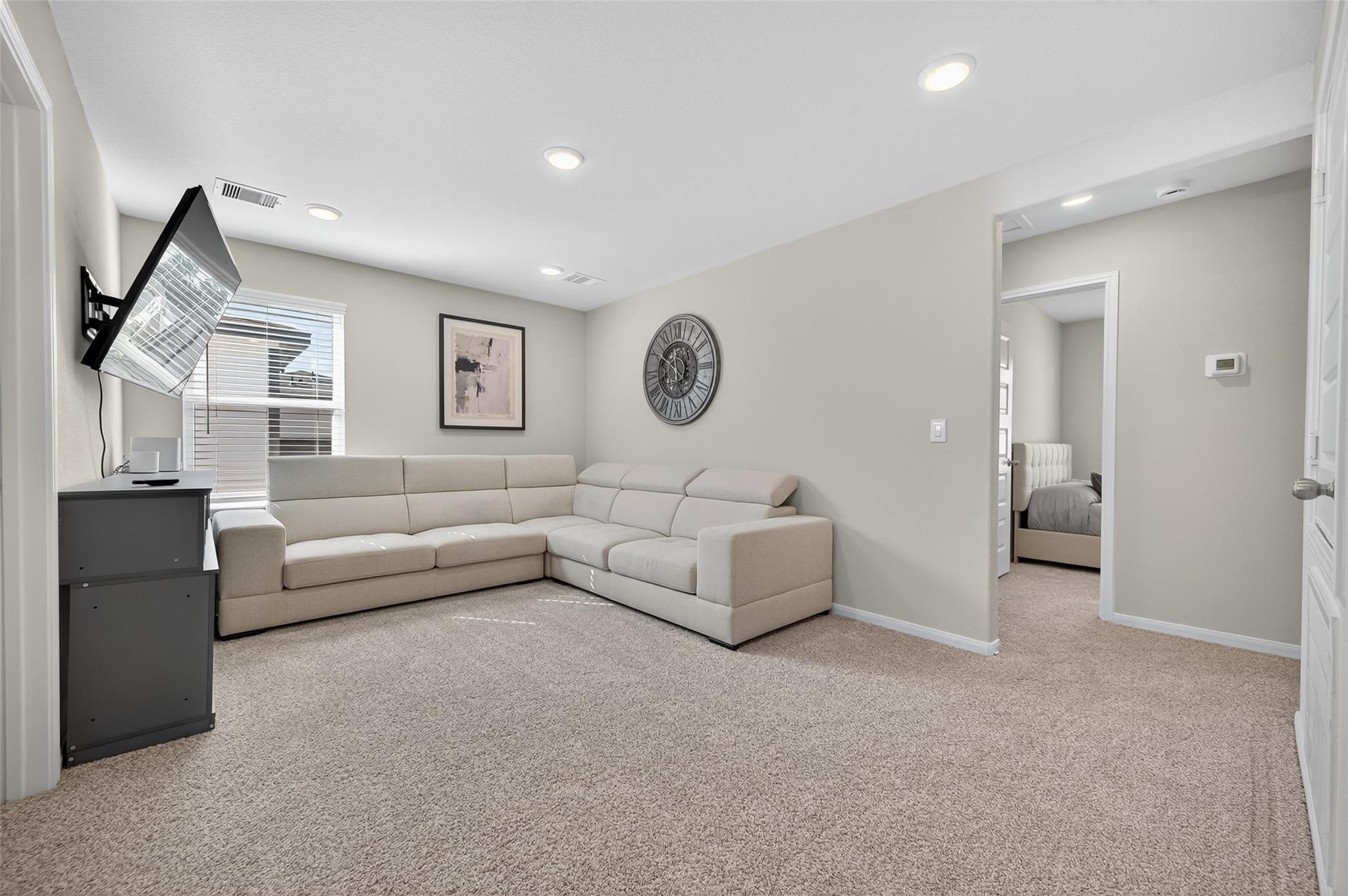 Cozy upstairs loft with beige L-shaped sofa, wall-mounted TV, round clock, and adjacent bedroom in Davidson Homes The Brazos E, Magnolia TX
