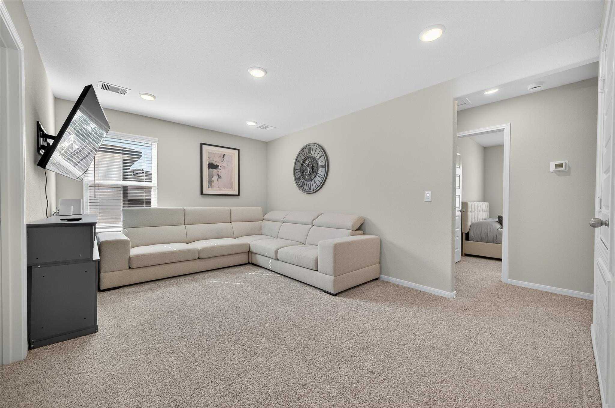 Cozy upstairs loft with beige L-shaped sofa, wall-mounted TV, round clock, and adjacent bedroom in Davidson Homes The Brazos E, Magnolia TX