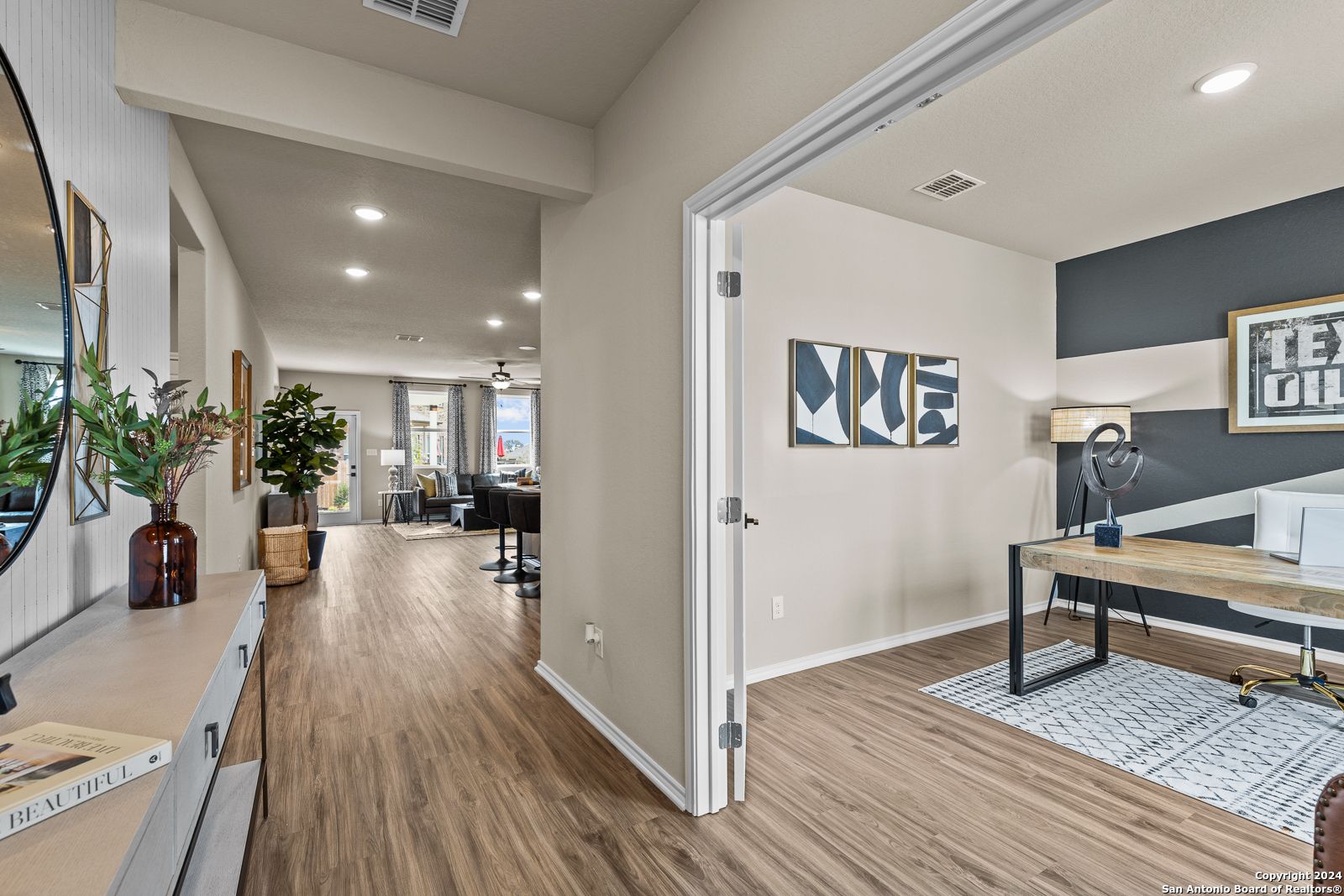 Spacious hallway with hardwood floors leading to modern home office, dark accent wall, wooden desk, and Texas art in Davidson Homes The Douglas F, Bricewood, San Antonio