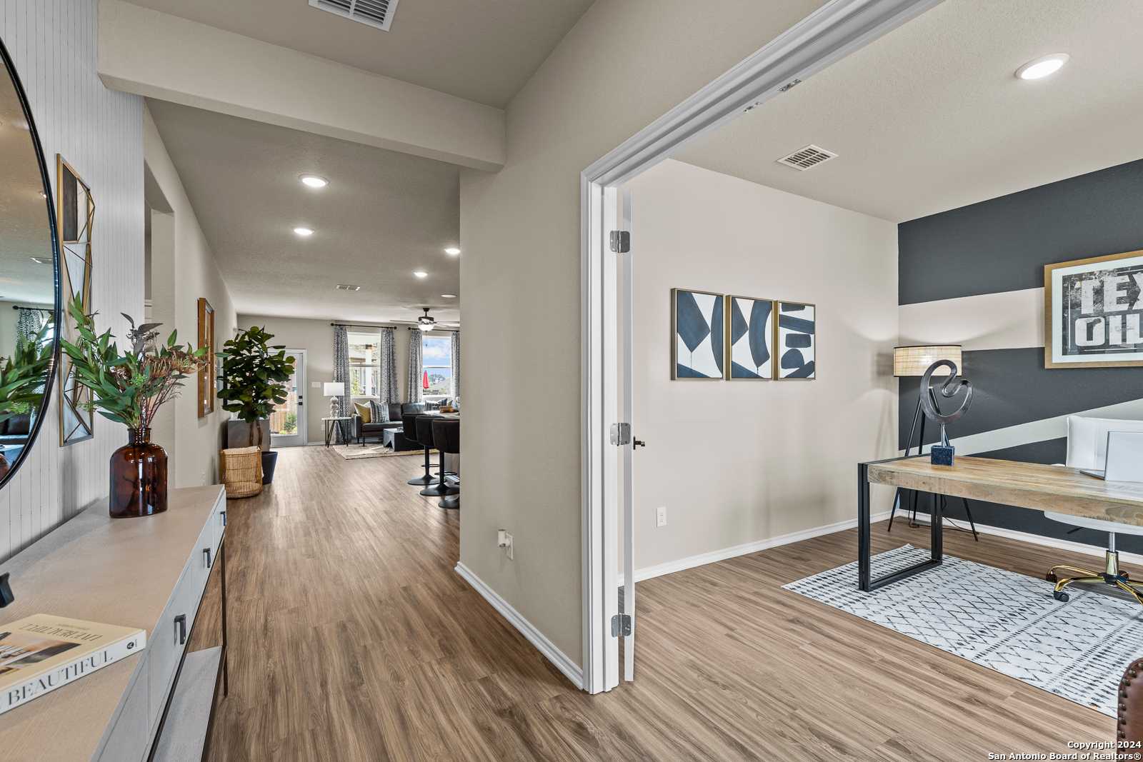 Spacious hallway with hardwood floors leading to modern home office, dark accent wall, wooden desk, and Texas art in Davidson Homes The Douglas F, Bricewood, San Antonio