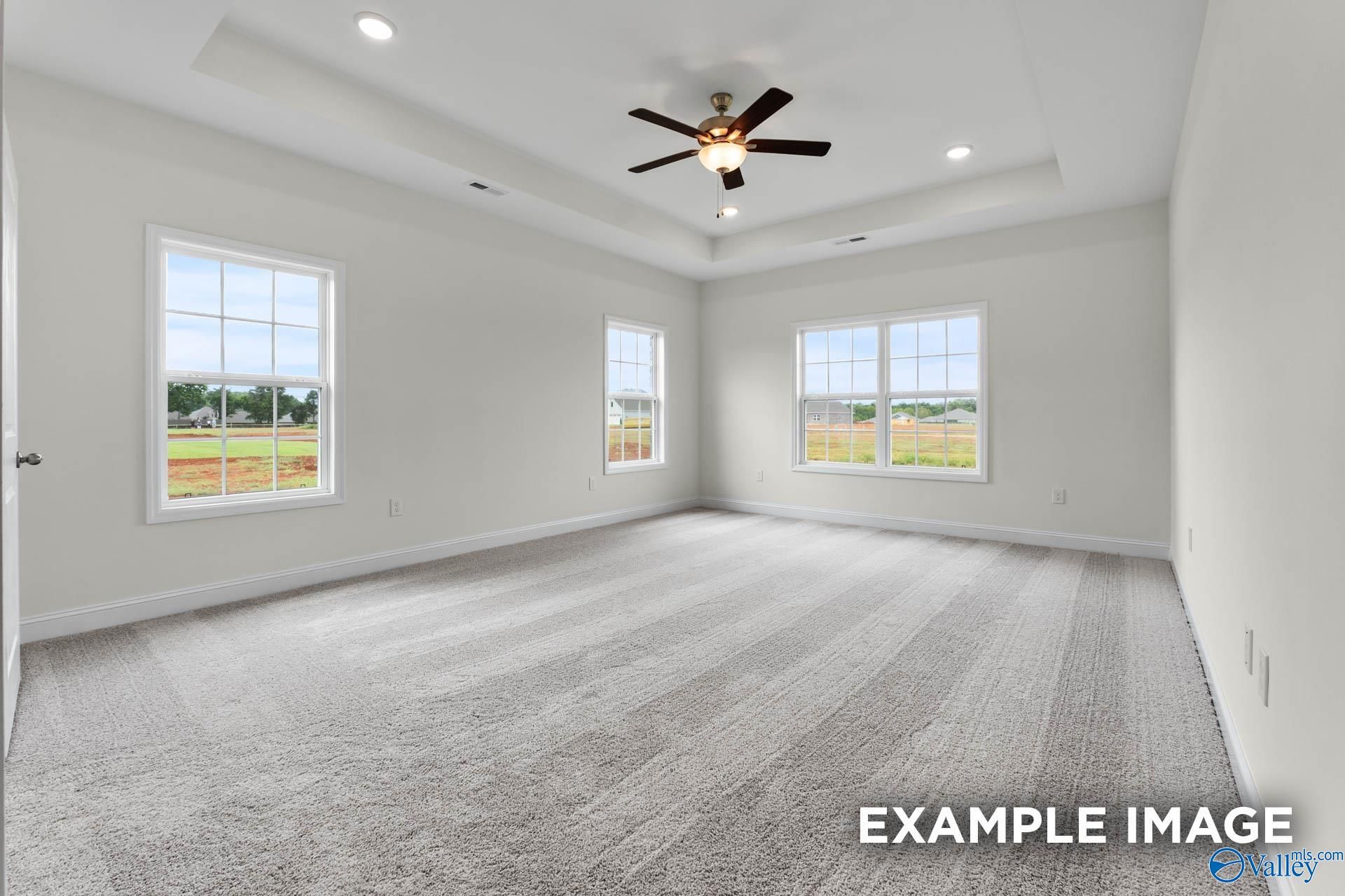 Spacious empty bedroom with gray carpet, large windows overlooking fields, tray ceiling fan in Davidson Homes The Emory, Toney, Alabama