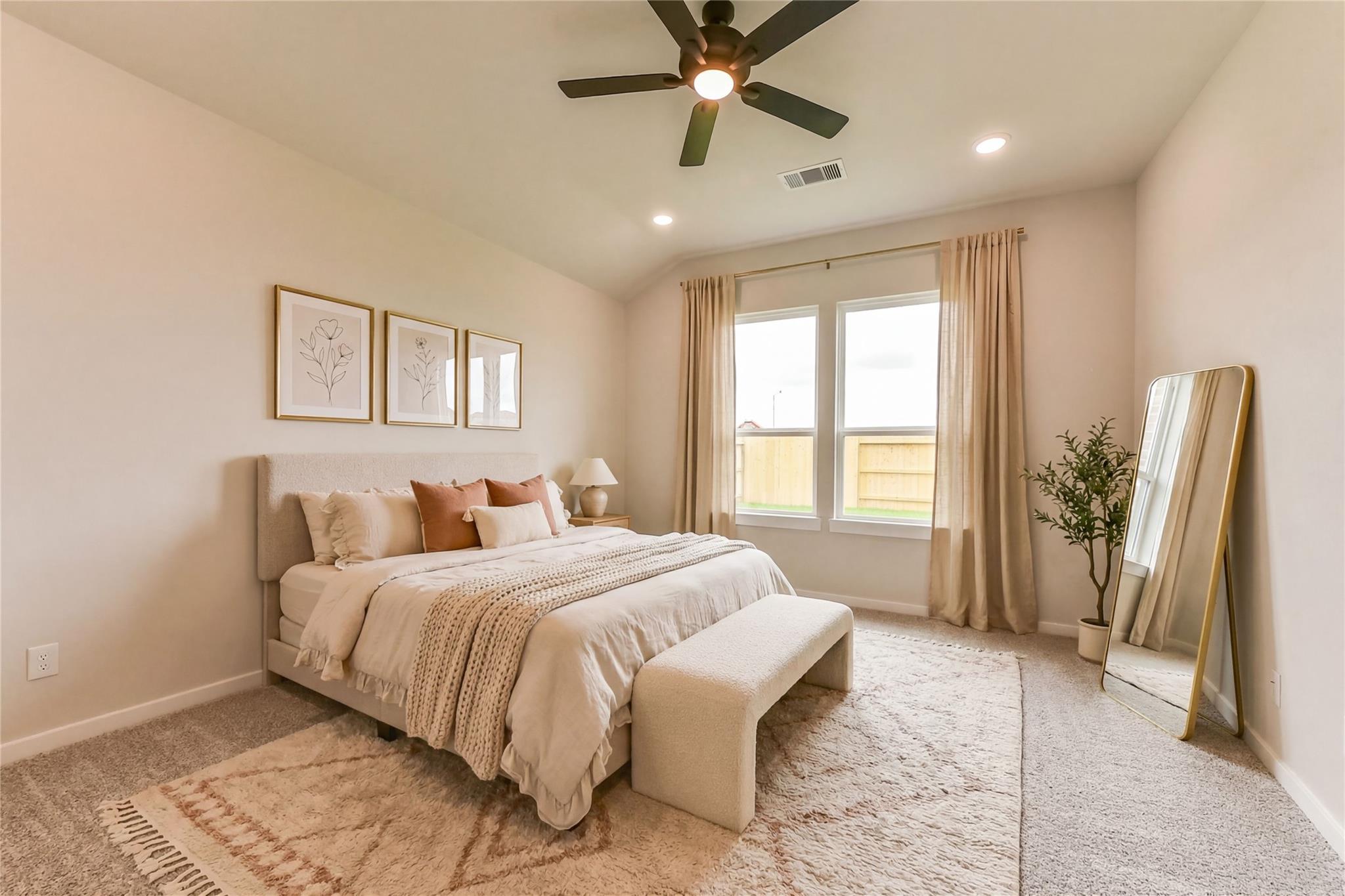 Elegant master bedroom with king bed, plush beige linens, ceiling fan, large windows, potted plant, and gold mirror in Davidson Homes Sequoia C, Crosby TX