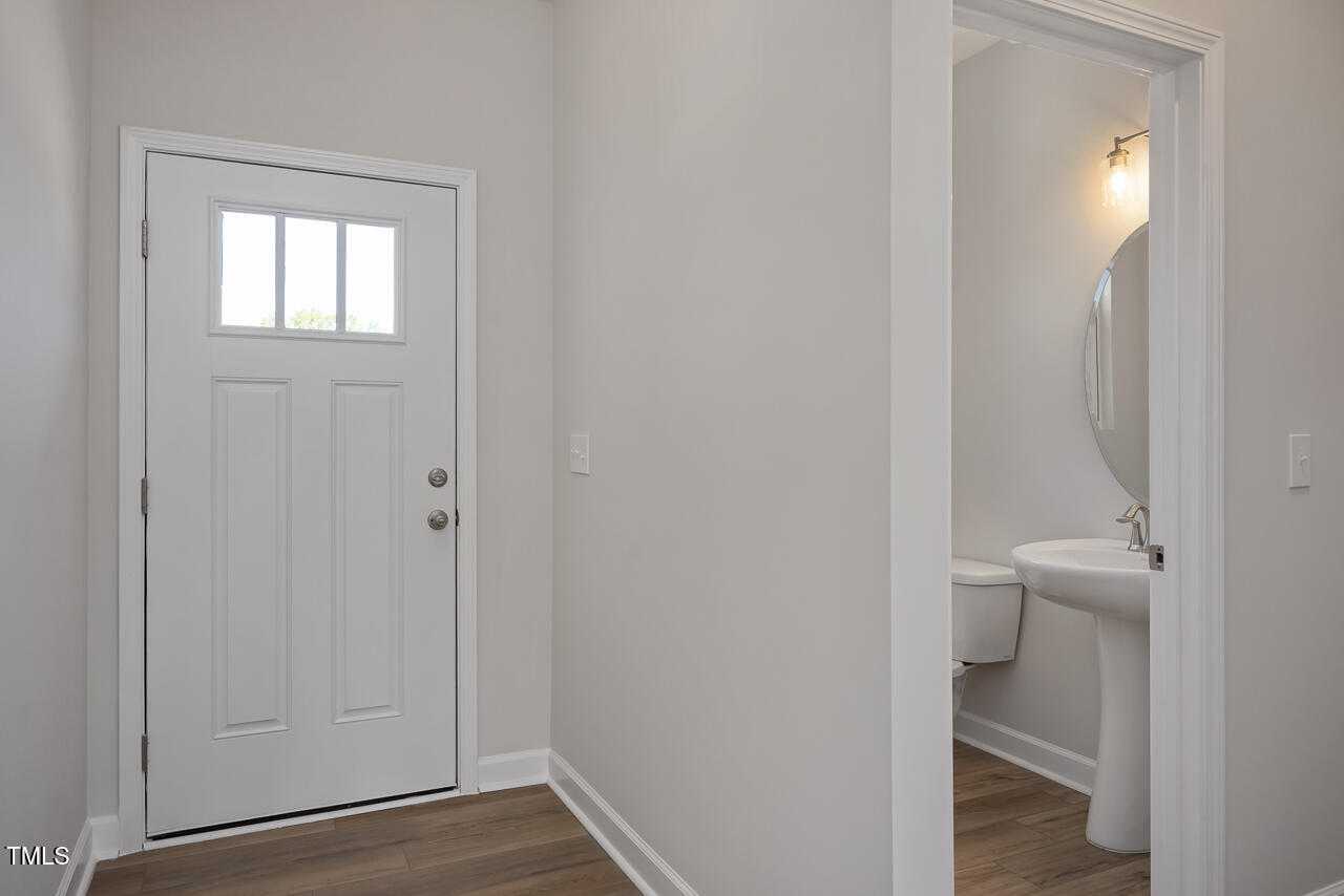 Bright entryway with white paneled door and adjacent powder room featuring pedestal sink, round mirror in Davidson Homes The Graham, Fuquay-Varina