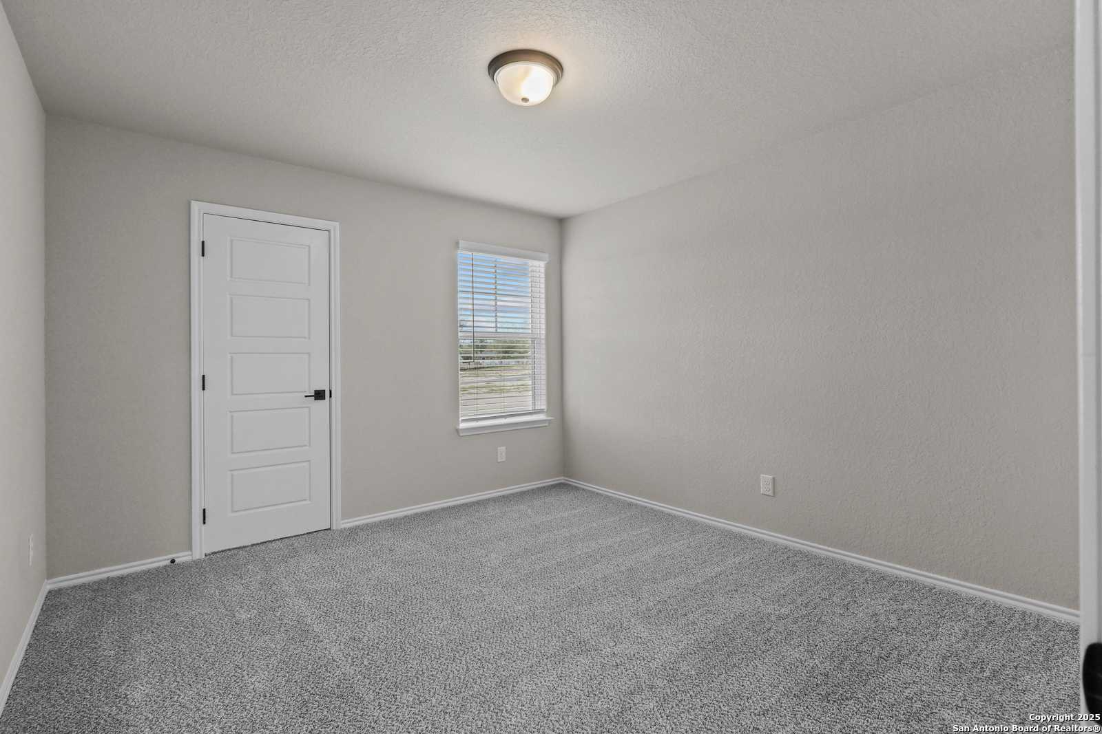 Spacious secondary bedroom with neutral gray walls, beige carpet, white door, and window blinds in Davidson Homes The Charlotte B, San Antonio