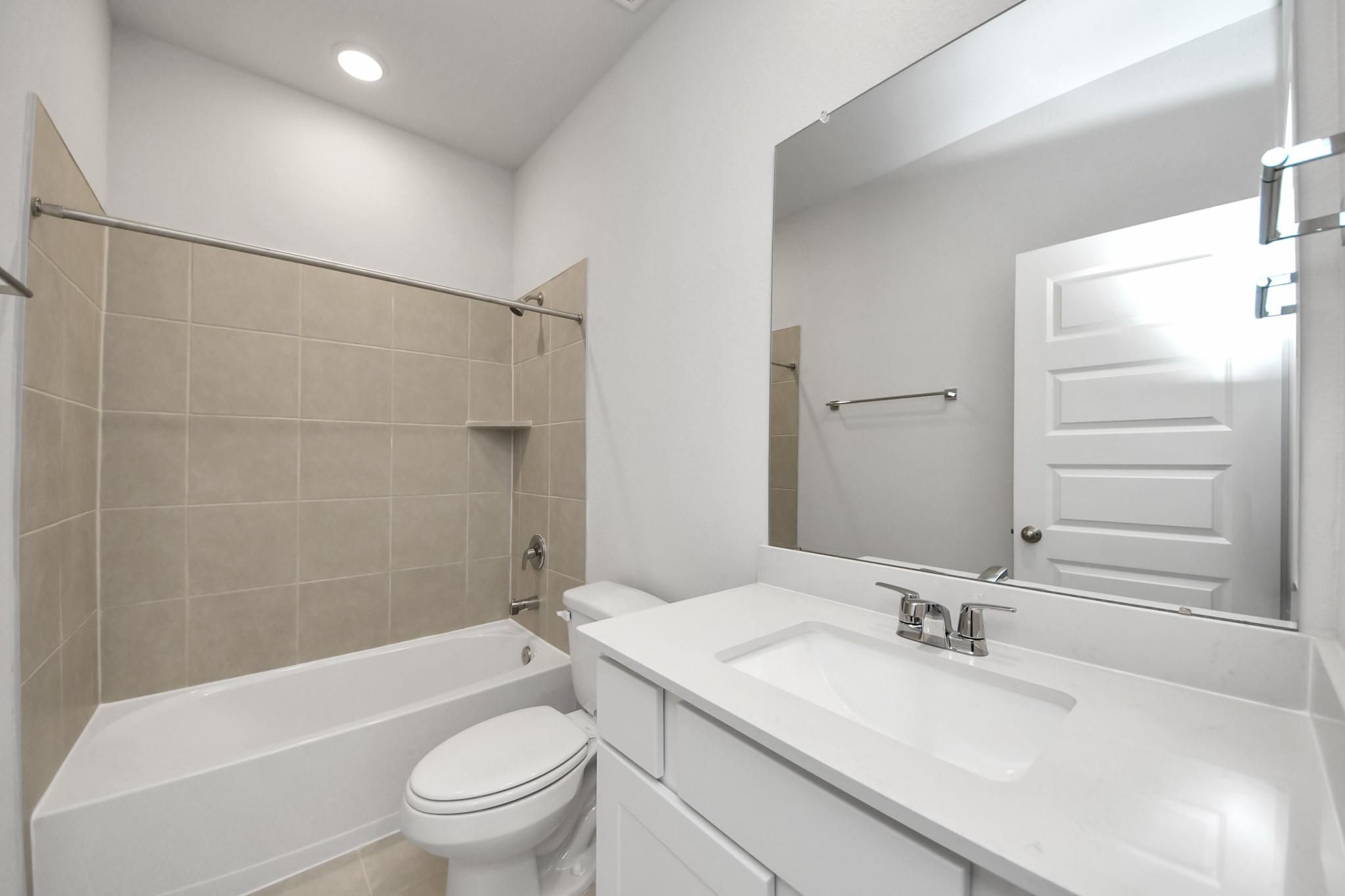 Bright white bathroom with tile tub-shower, toilet, and large vanity mirror in Davidson Homes The Costa A, Emberly, Beasley, Texas