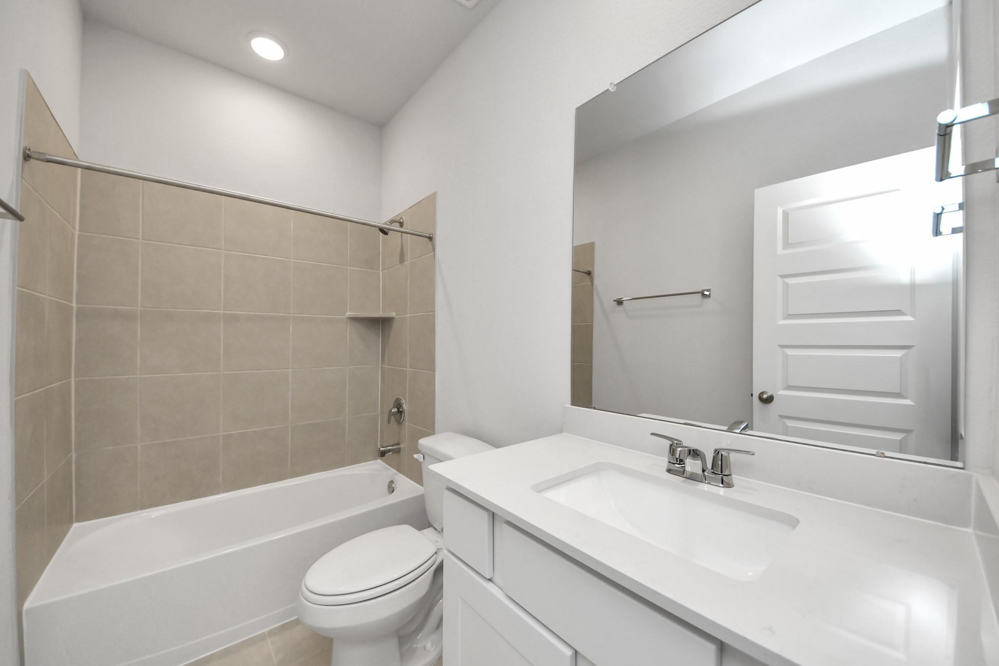 Bright white bathroom with tile tub-shower, toilet, and large vanity mirror in Davidson Homes The Costa A, Emberly, Beasley, Texas