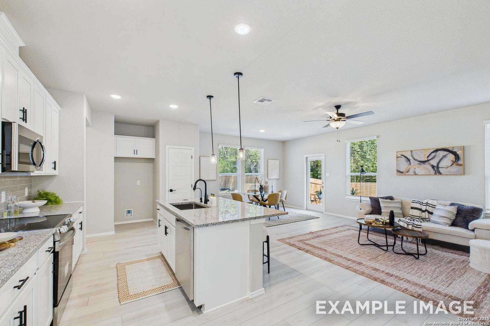 Open-concept kitchen and living area with white cabinets, island bar, ceiling fan, and backyard views in Davidson Homes The Charlotte B, San Antonio