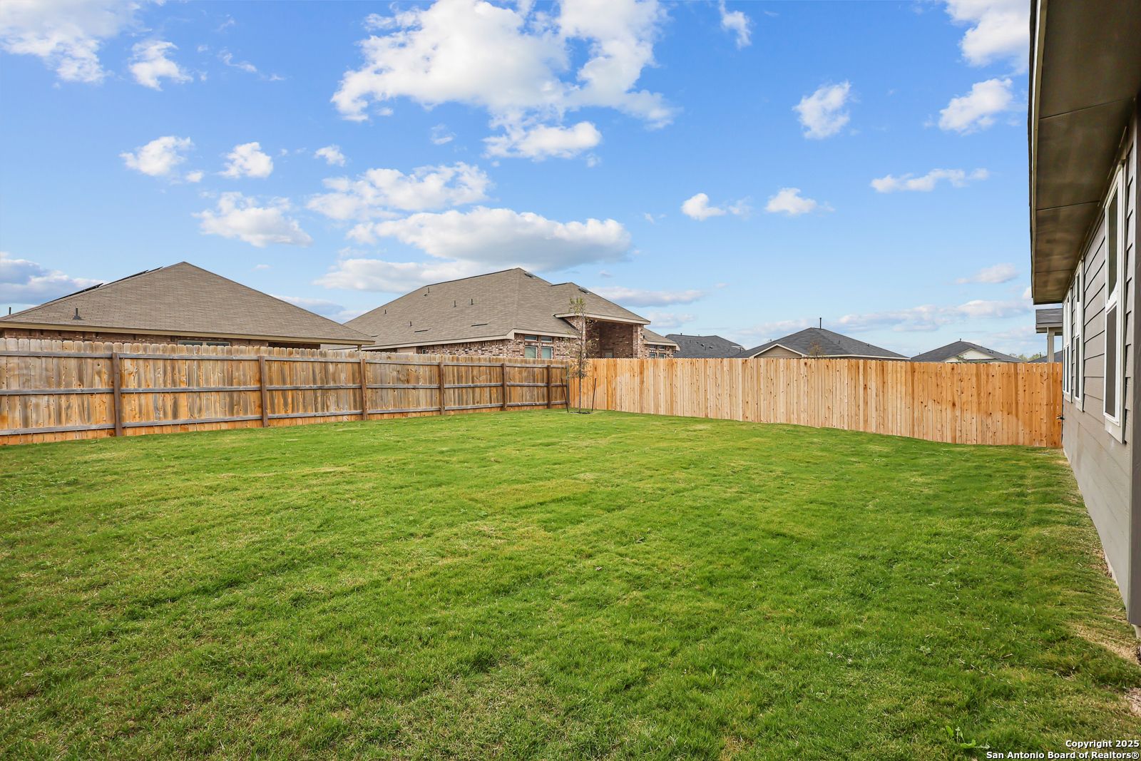 Lush green backyard with wooden privacy fence and neighboring homes under blue sky in Davidson Homes The Daphne J, Converse Texas