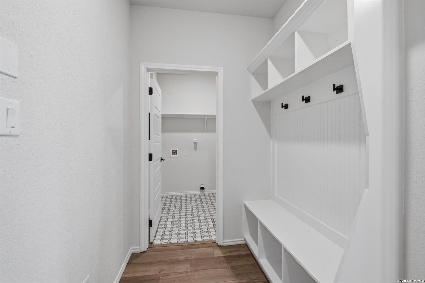White mudroom featuring built-in bench, shelves, coat hooks, and open laundry room with patterned tile in Davidson Homes The Blanco C, San Antonio