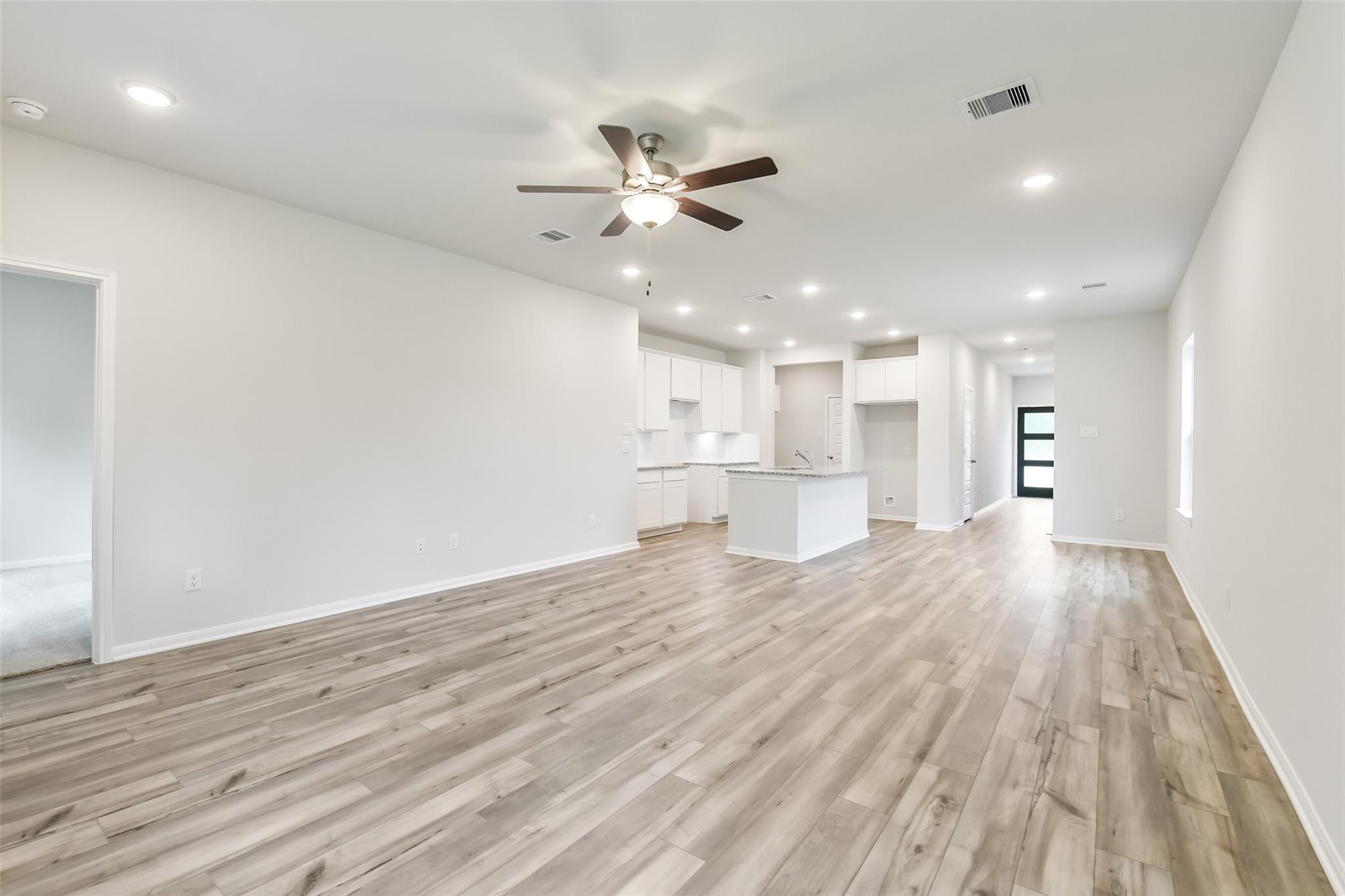 Spacious open living area with kitchen island, white cabinets, ceiling fan, and light hardwood floors in The Brazos F home, Conroe, Texas