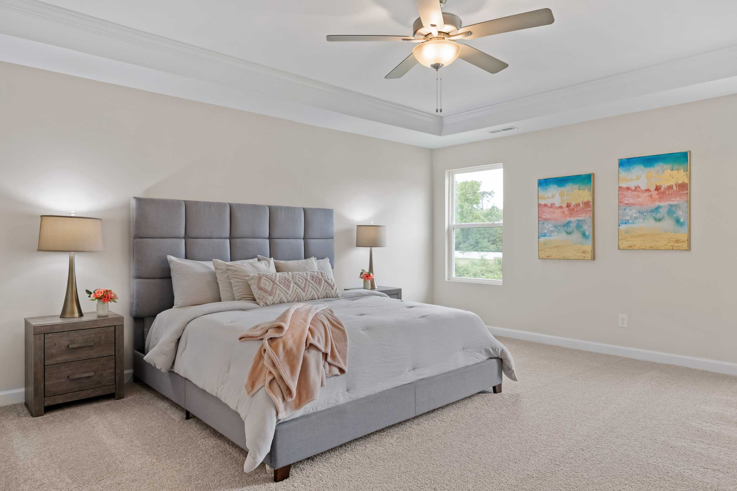 Spacious primary bedroom at Gregory Village in Lillington NC with gray upholstered king bed, neutral tones, ceiling fan, and abstract wall art