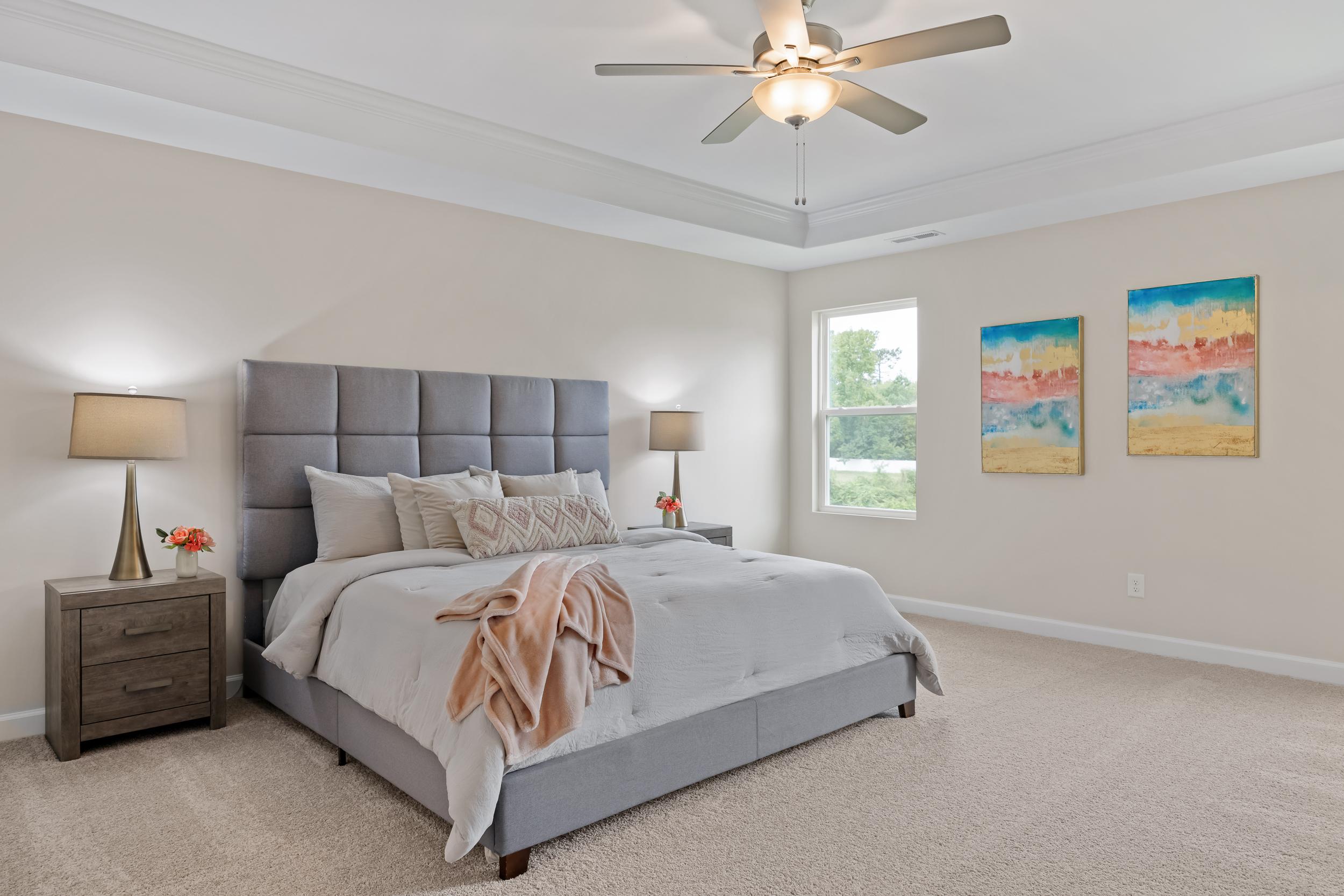 Spacious primary bedroom at Gregory Village in Lillington NC with gray upholstered king bed, neutral tones, ceiling fan, and abstract wall art