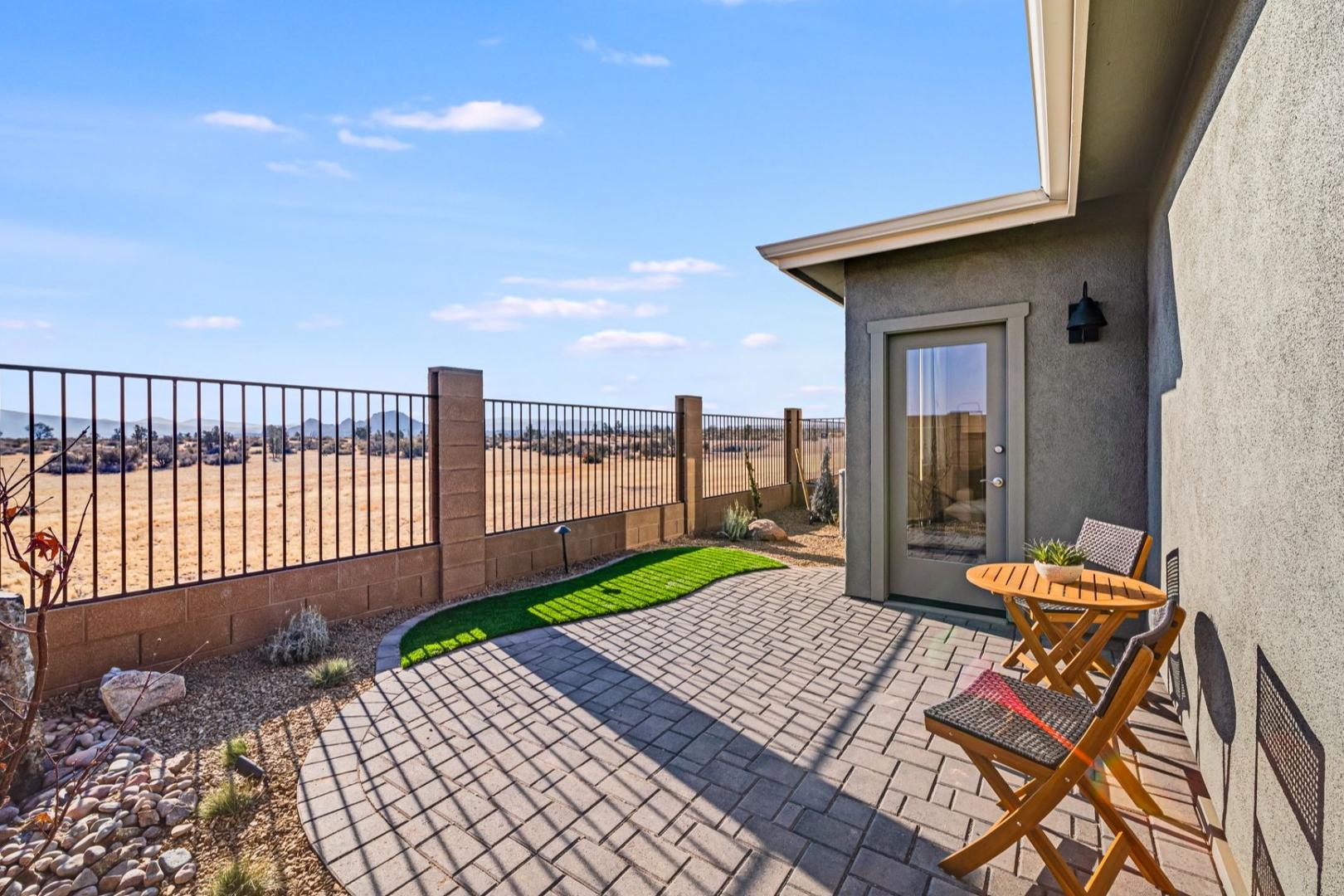 Spacious desert patio of The Savannah B home in Prescott, AZ, with wooden chairs, small table, sliding glass door, paver tiles, and metal fence overlooking arid landscape